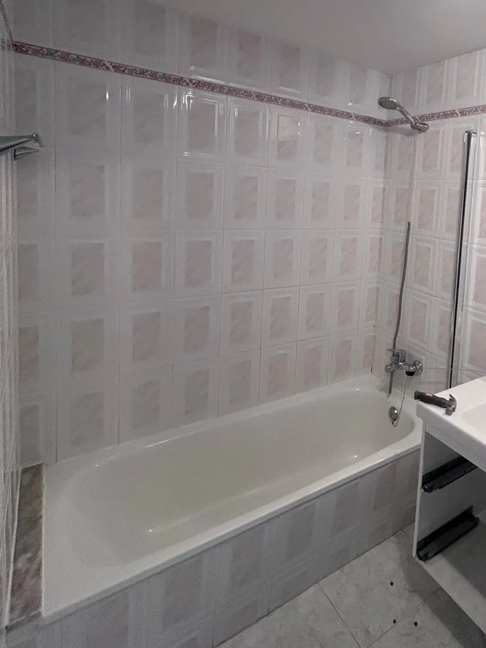 Bany amb banyera abans de l’obra: banyera blanca amb el faldó enrajolat, aixeta amb flexible i parets de rajola clara amb quadrats rosats i sanefa de flors; a la dreta s’hi veu la cantonada del lavabo