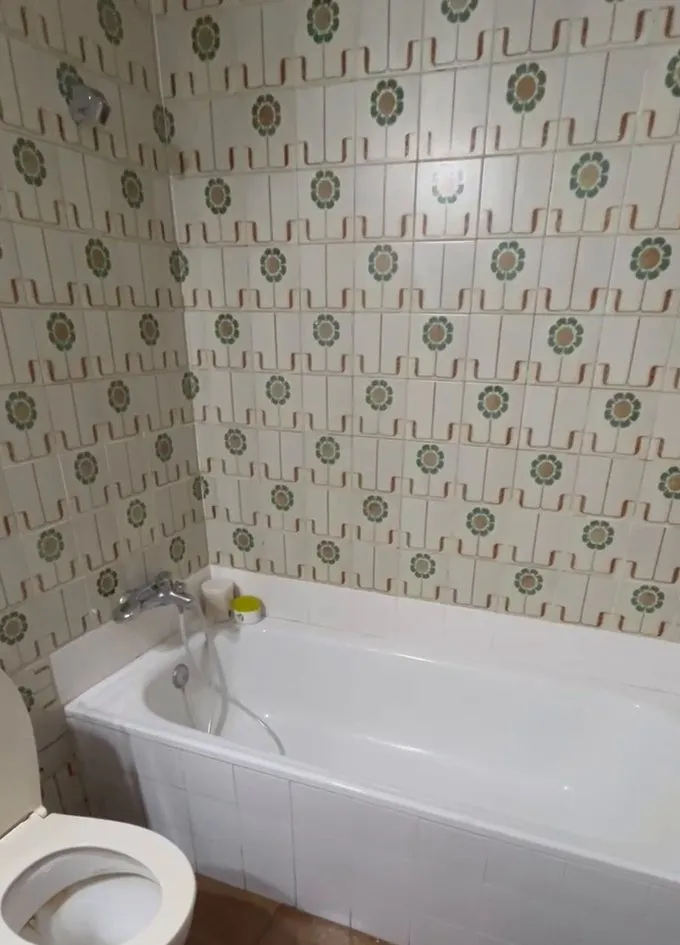 Bañera blanca con el faldón alicatado en blanco y el grifo con flexo, bajo paredes de azulejo antiguo de flores verdes y ocres; a la izquierda, el inodoro