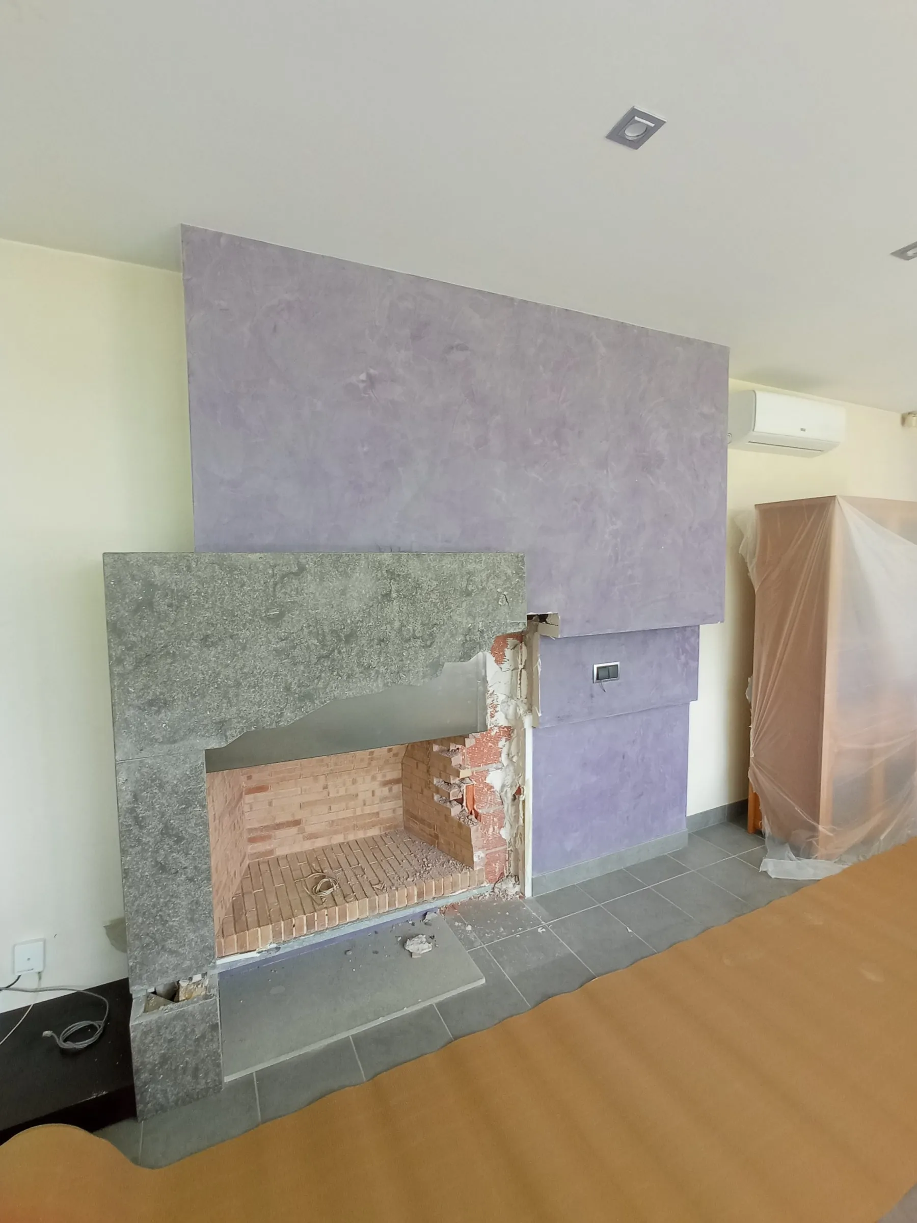 Chimenea con hogar de ladrillo y marco de piedra gris, en una pared lila de acabado texturado; el suelo está protegido con cartón, a la derecha hay un mueble cubierto con plástico y ya se ven algunas piezas rotas