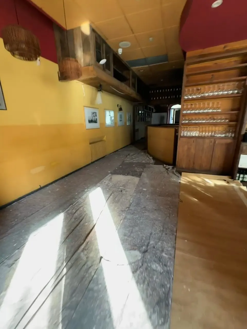 El vestíbul de Ca la Paula abans de la reforma: parets pintades de groc amb petits quadres emmarcats, prestatgeria de fusta amb copes, sostre amb un tram vermell i terra de fusta fosca cobert en part amb paper de protecció, amb franges de sol