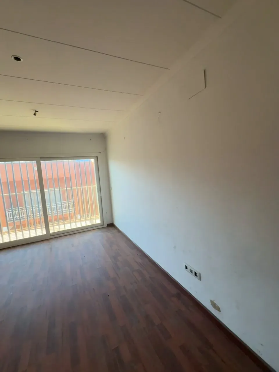 Salón antes de la reforma, con suelo de lamas oscuras, techo de placas blancas y una puerta corredera al balcón