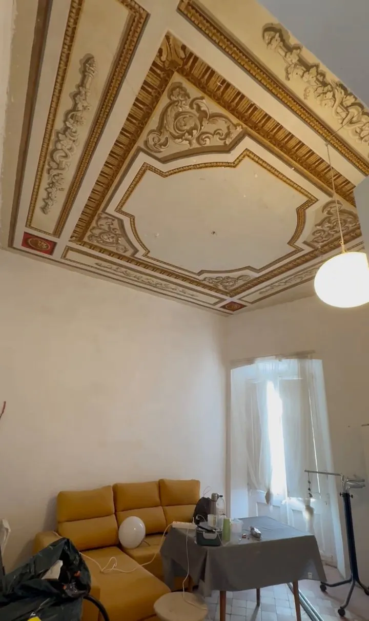 Salón antes de reformarlo, con el techo decorado con molduras y motivos pintados en dorado y crema, un sofá mostaza, una mesa con mantel gris y una bolsa negra en primer plano