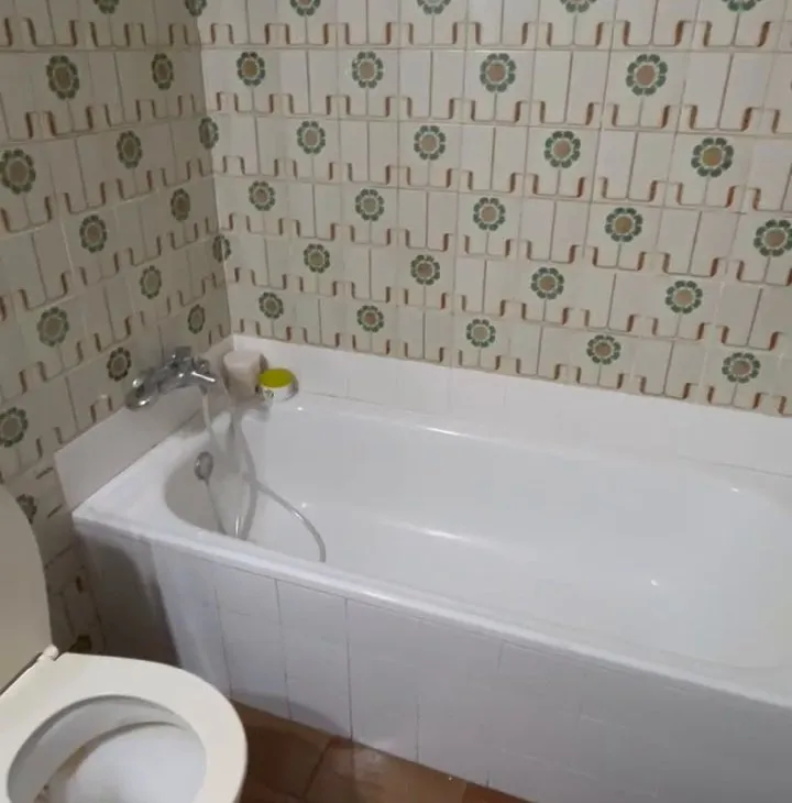 Vista cercana de la bañera blanca con el faldón alicatado en blanco y el grifo con flexo, junto a la pared de azulejo antiguo; abajo a la izquierda, el inodoro