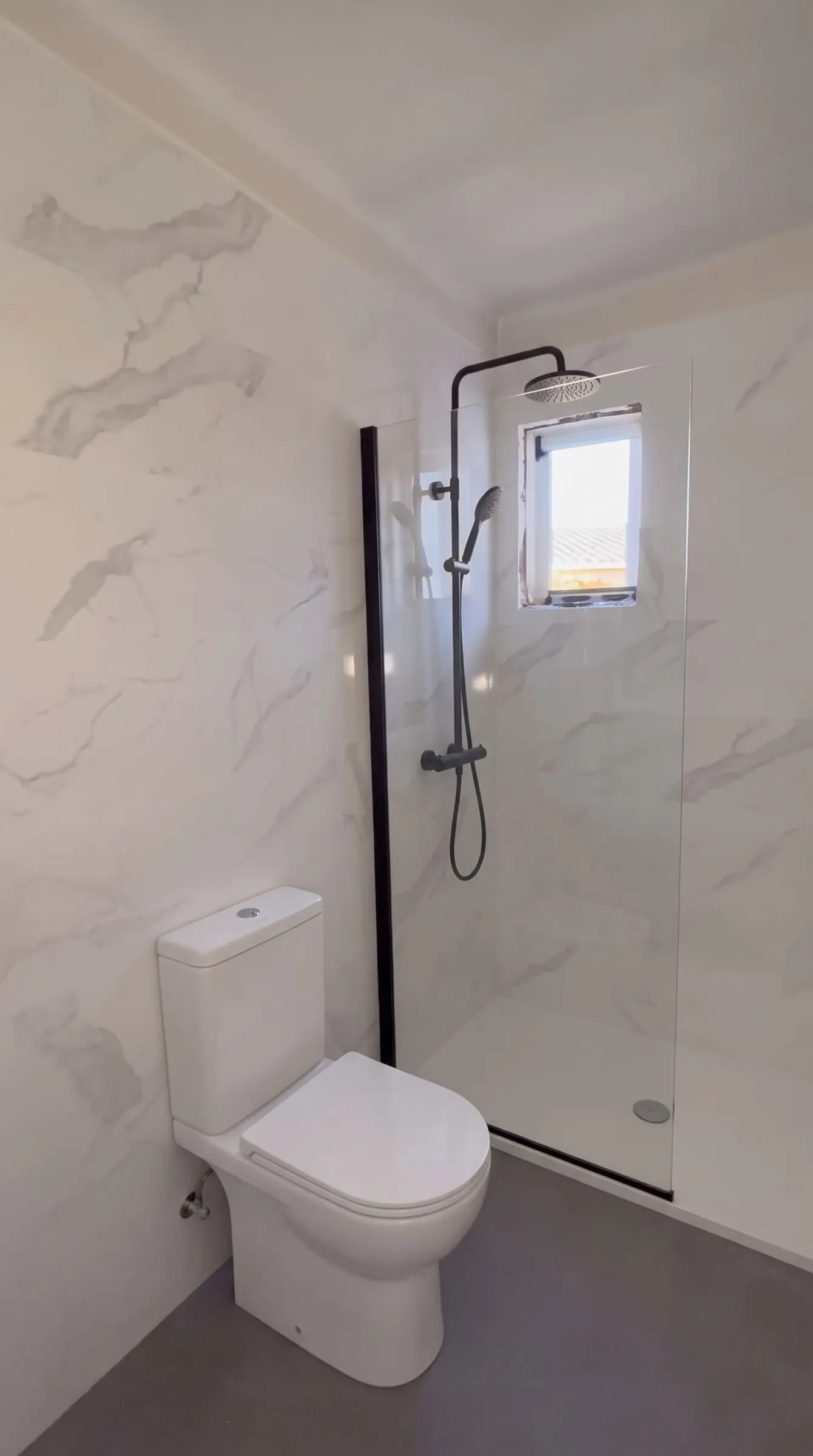 Baño de Banyoles con paredes de baldosa blanca de aspecto marmóreo y suelo gris oscuro, con un inodoro, una columna de ducha y una mampara de vidrio añadidos con ambientación digital sobre un fotograma de vídeo de la obra