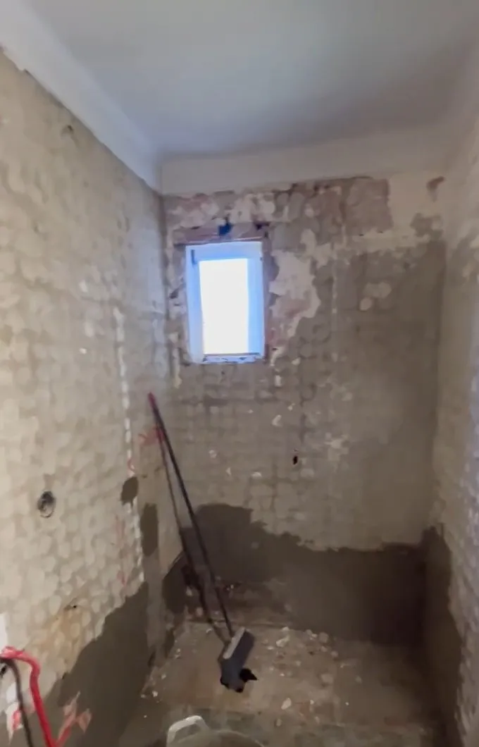 Baño en obra con las paredes sin alicatado y con restos de mortero, una ventana pequeña, una franja oscura en la parte baja de las paredes y un cubo y herramientas en el suelo