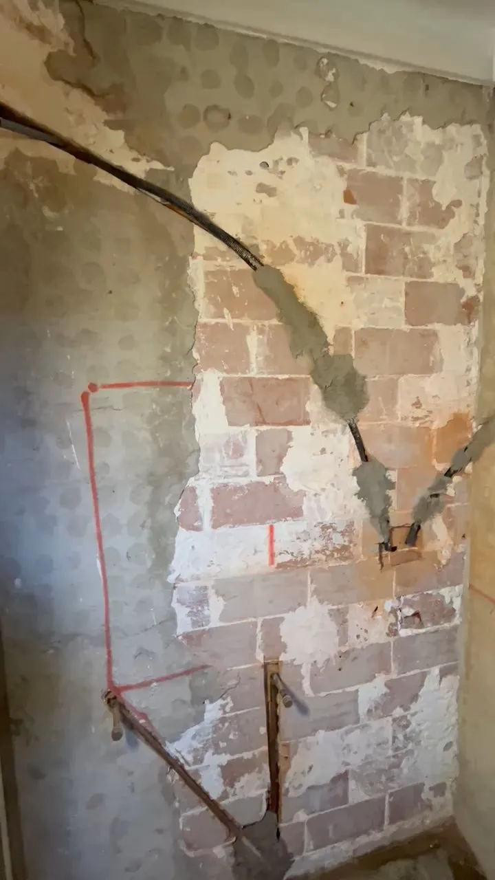 Pared de ladrillo al descubierto con rozas, un tubo negro empotrado y tubos metálicos en la parte baja, durante el derribo de un baño
