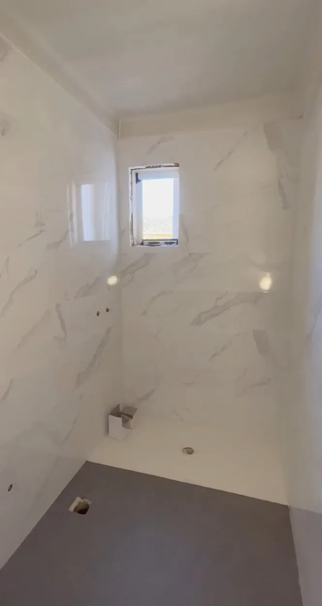 Baño con las paredes alicatadas con azulejo blanco de aspecto marmóreo, ventana pequeña, base blanca con desagüe y suelo gris oscuro, aún sin sanitarios