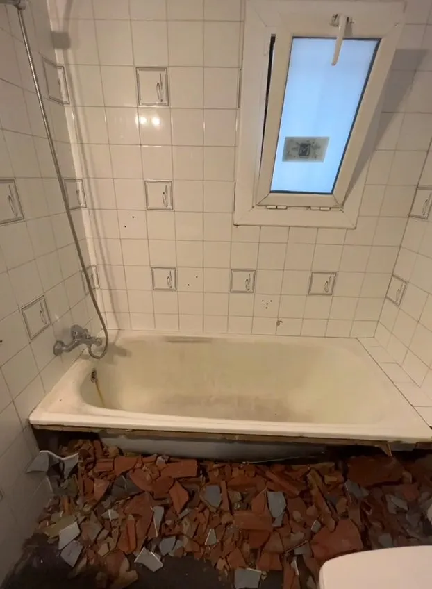 Baño con una bañera de color amarillento y azulejos blancos con piezas decorativas; restos de ladrillo y azulejo roto en el suelo, delante de la bañera