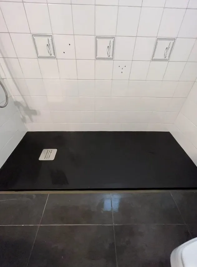 Plato de ducha negro con rejilla metálica de desagüe junto a una pared de azulejos blancos con piezas decorativas; delante, suelo de baldosa oscura y brillante
