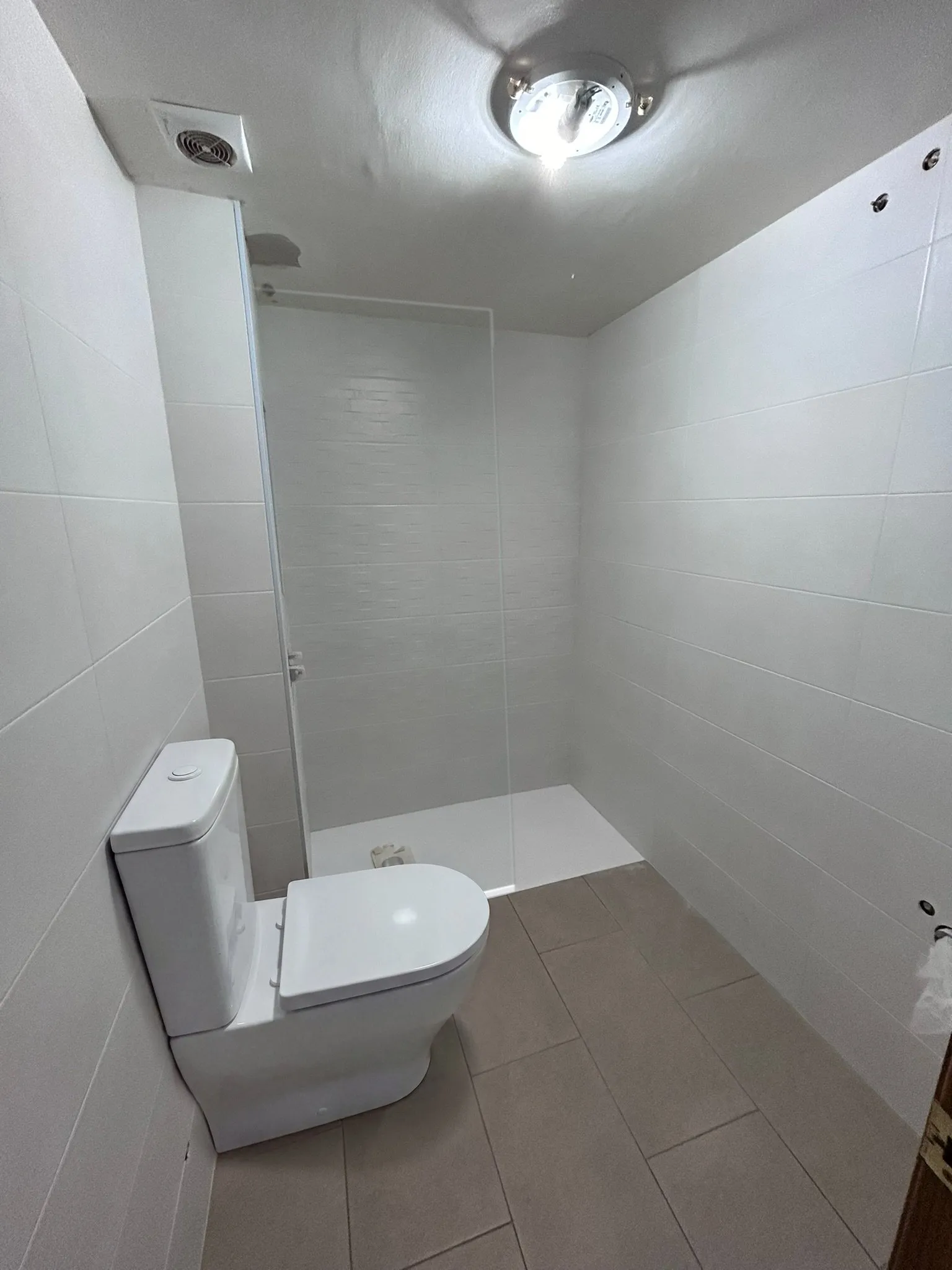 Baño en obra de una reforma completa en Banyoles, con las paredes alicatadas en blanco, el plato de ducha blanco y el suelo de baldosa rectangular gris pardo ya colocados. Con ambientación digital sobre una fotografía de la obra se han añadido un inodoro blanco de suelo con cisterna y una mampara de vidrio.