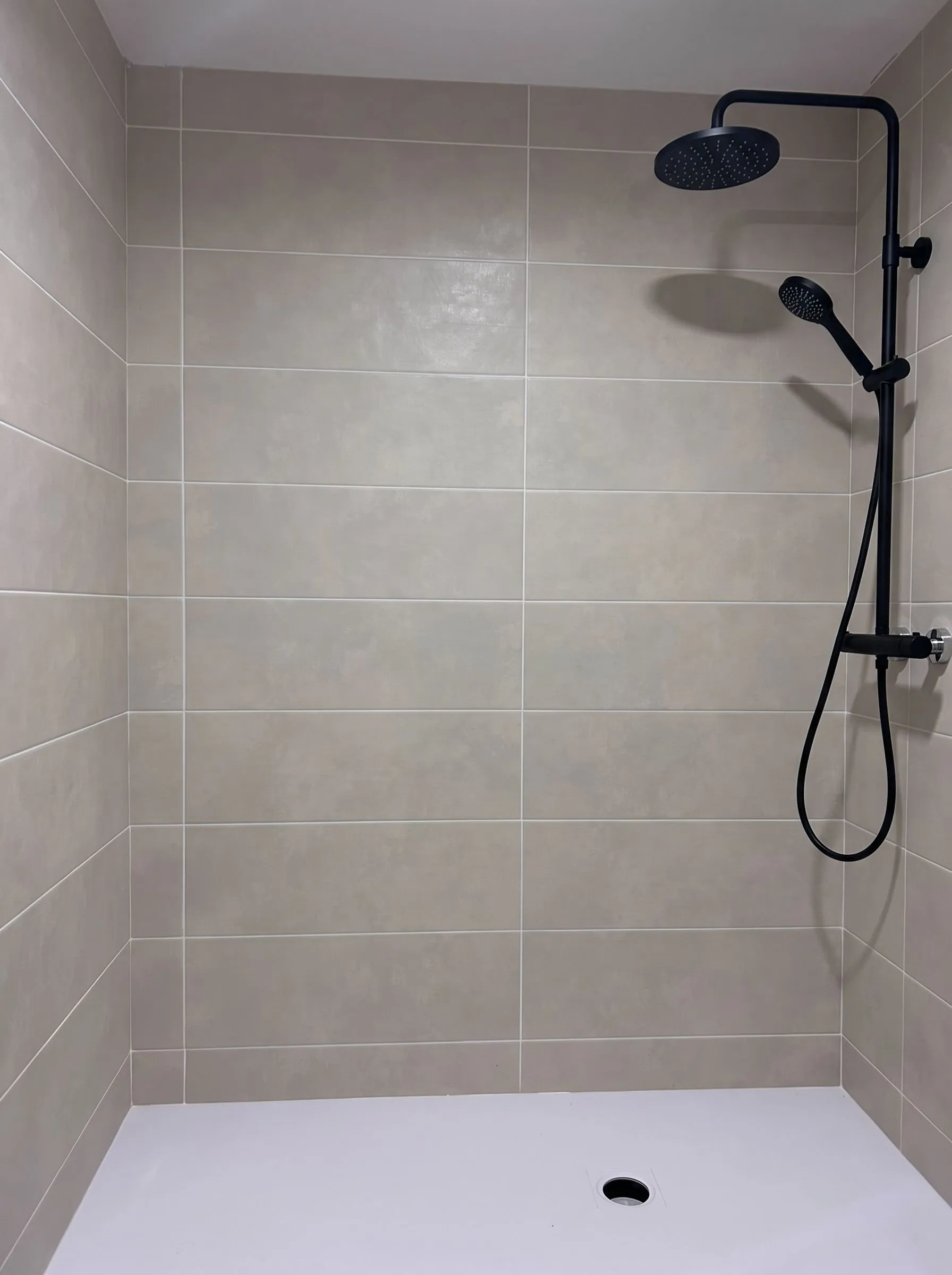 Ducha de un baño de Banyoles donde se cambió la bañera por un plato de ducha blanco, con las paredes alicatadas con azulejo rectangular gris claro y juntas blancas. Con ambientación digital sobre una fotografía de la obra se ha añadido una columna de ducha negra, con rociador redondo, ducha de mano y flexible, sobre las dos piezas cromadas que ya había en la pared derecha. No se ha añadido mampara.