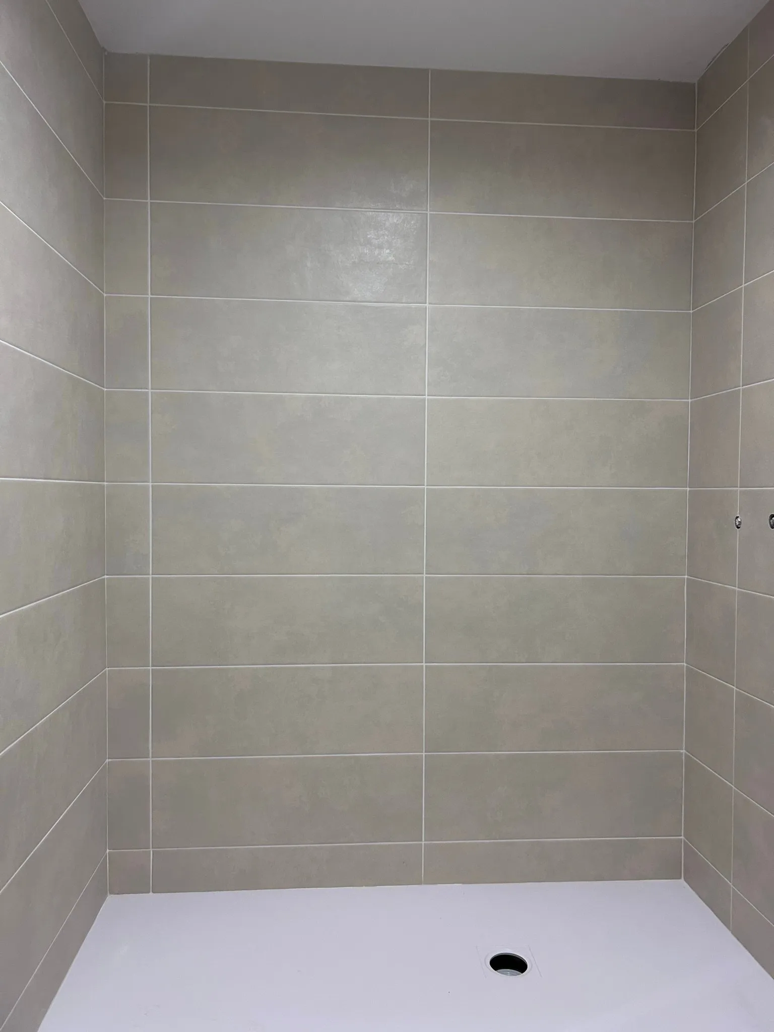 Ducha con las paredes alicatadas con azulejo rectangular gris claro y juntas blancas, y un plato de ducha blanco con el desagüe a la vista, aún sin grifería ni mampara