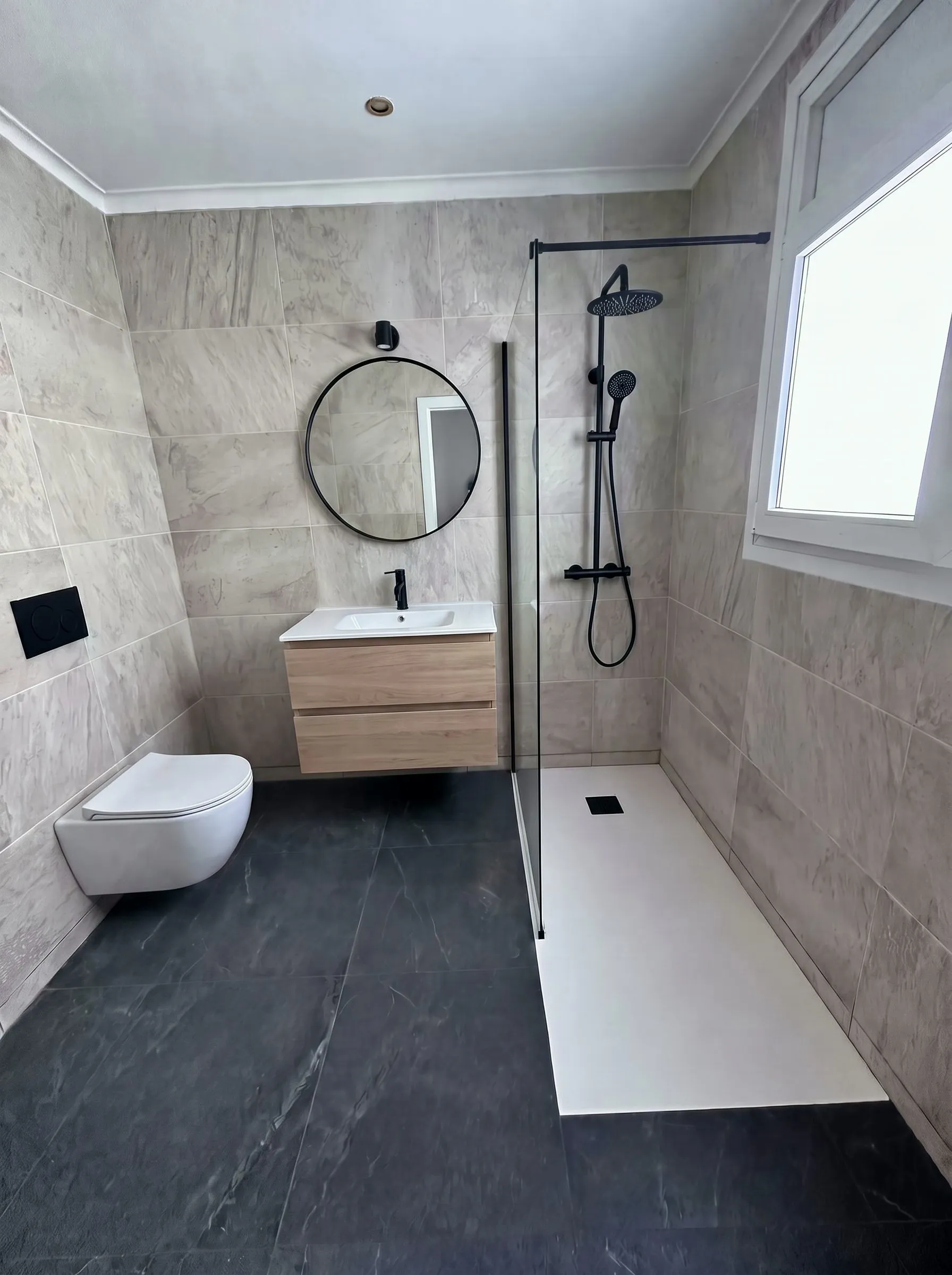 Baño reformado en Banyoles: columna de ducha negra, mampara y mueble suspendido de roble