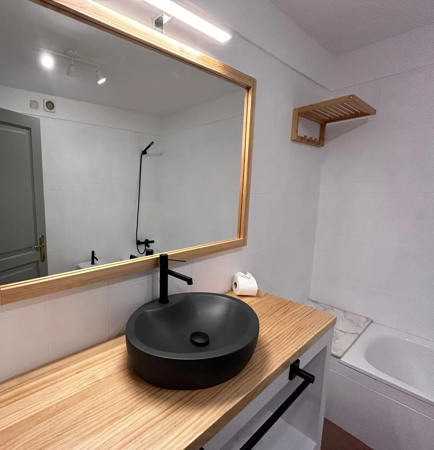 Baño reformado en Camallera: lavabo negro mate sobre encimera de madera clara, grifo negro alto, espejo grande con marco de madera y luz encima, paredes de azulejo blanco y, a la derecha, una bañera