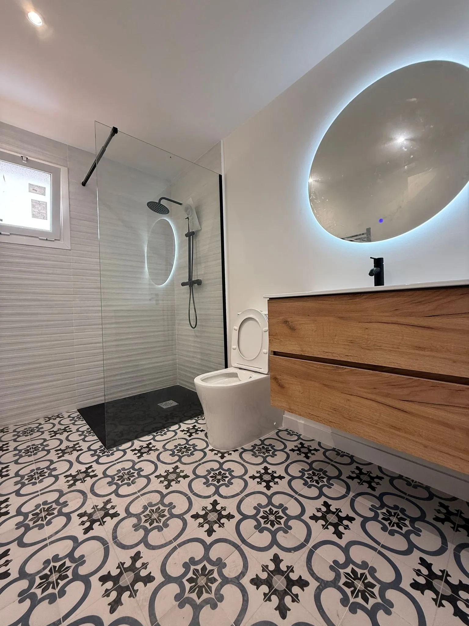 Baño reformado en Girona con suelo de dibujo hidráulico, ducha de plato negro y mueble de madera con espejo redondo iluminado
