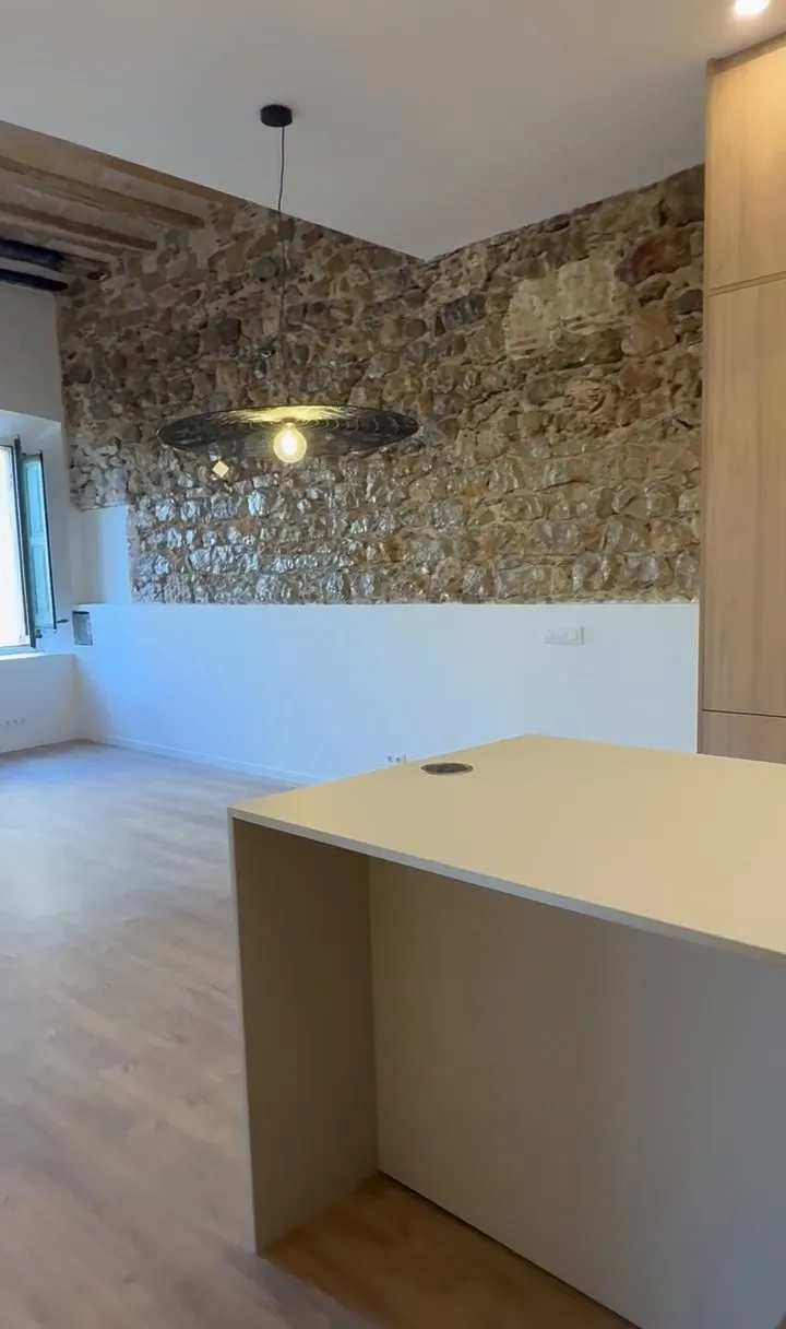 Cocina con isla de encimera clara junto a una pared de piedra vista, en un piso reformado en el Barri Vell de Girona