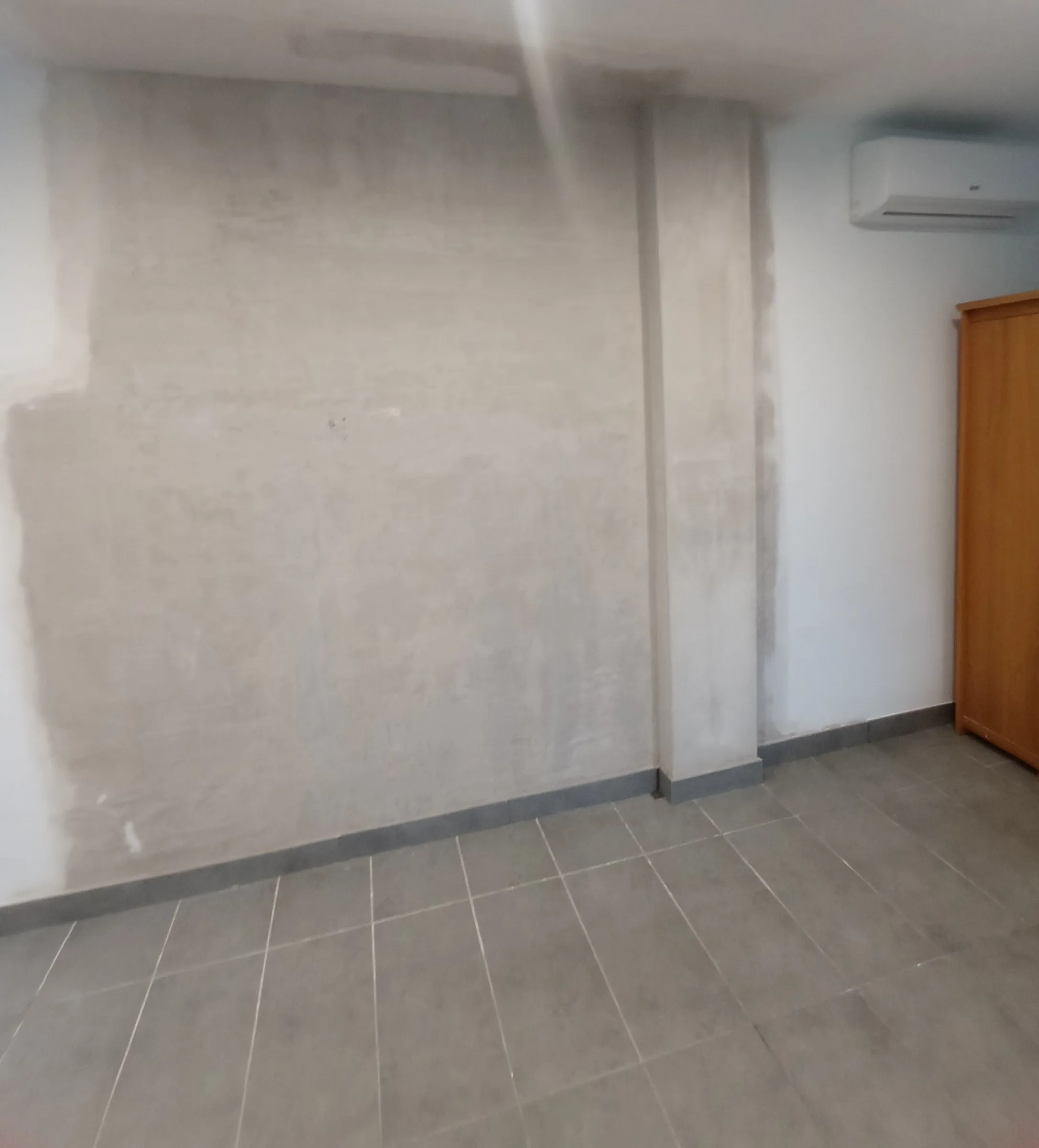 Estancia con una pared enyesada y lisa, de color gris claro, lista para pintar, con un resalte vertical a la derecha; suelo de baldosa gris con rodapié gris, un aparato de aire acondicionado y un armario de madera a la derecha