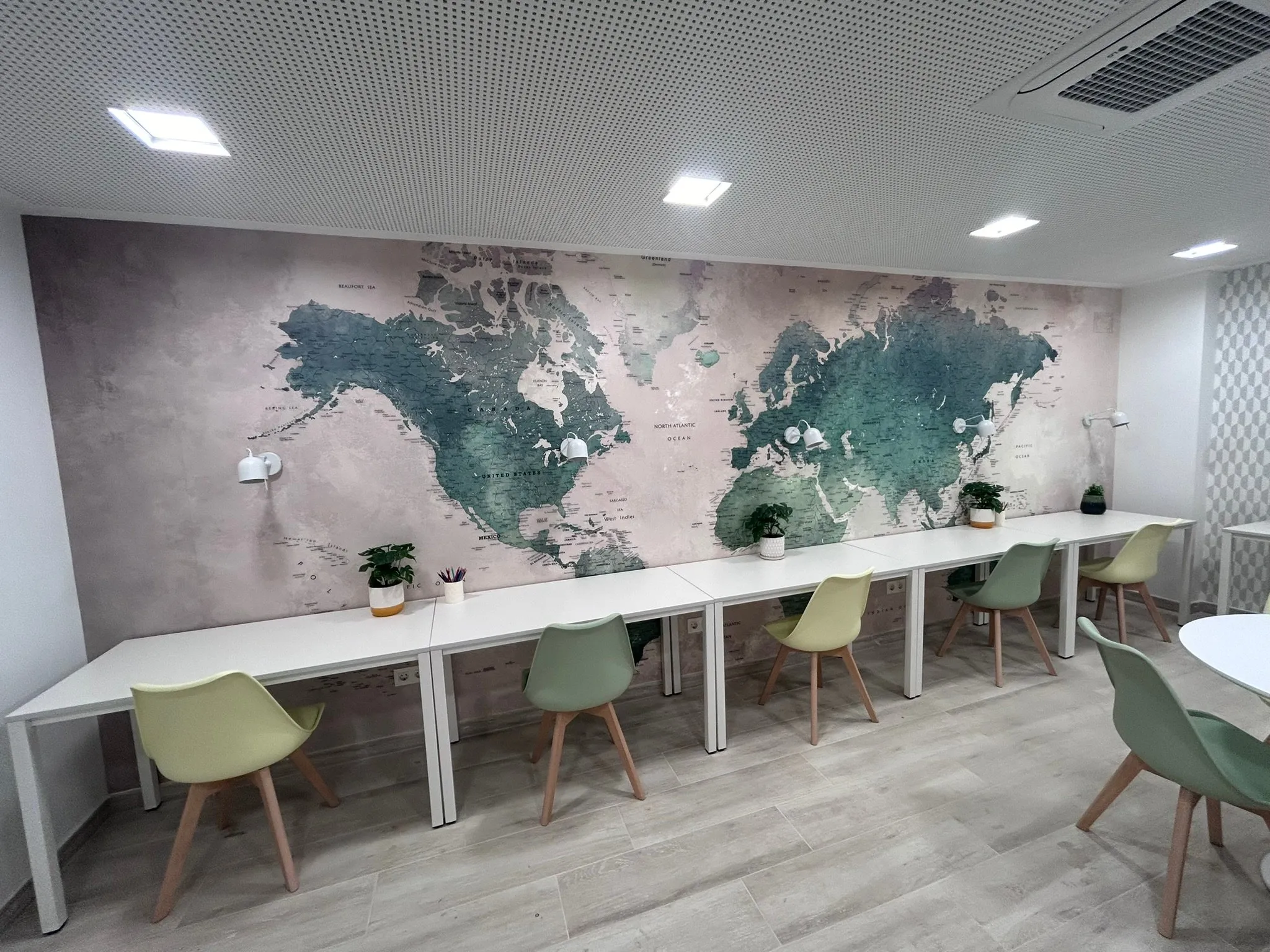 La sala del mapamundi, a la sala de jocs d’un bloc de pisos de Girona: un gran mural de mapamundi, taules llargues blanques amb cadires en tons verds i grocs, aplics de paret i fals sostre perforat amb focus