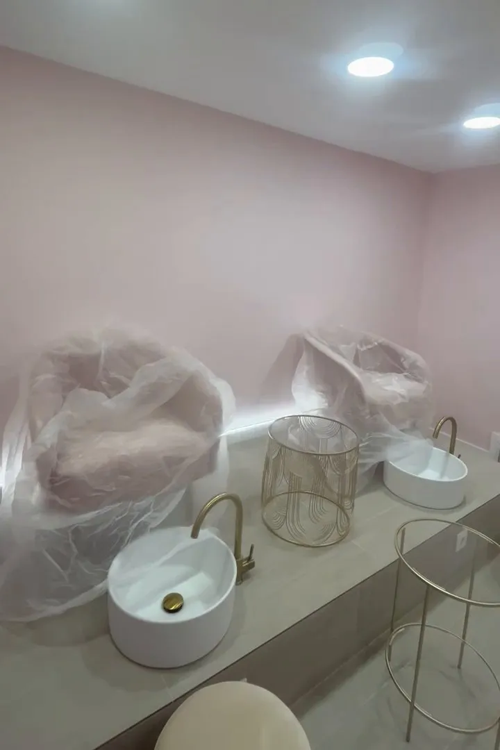 Sala amb parets rosa pàl·lid i focus al sostre; sobre una plataforma elevada d’acabat gris clar hi ha dues butaques cobertes amb plàstic, dos lavabos rodons blancs amb aixeta daurada i tauletes auxiliars daurades