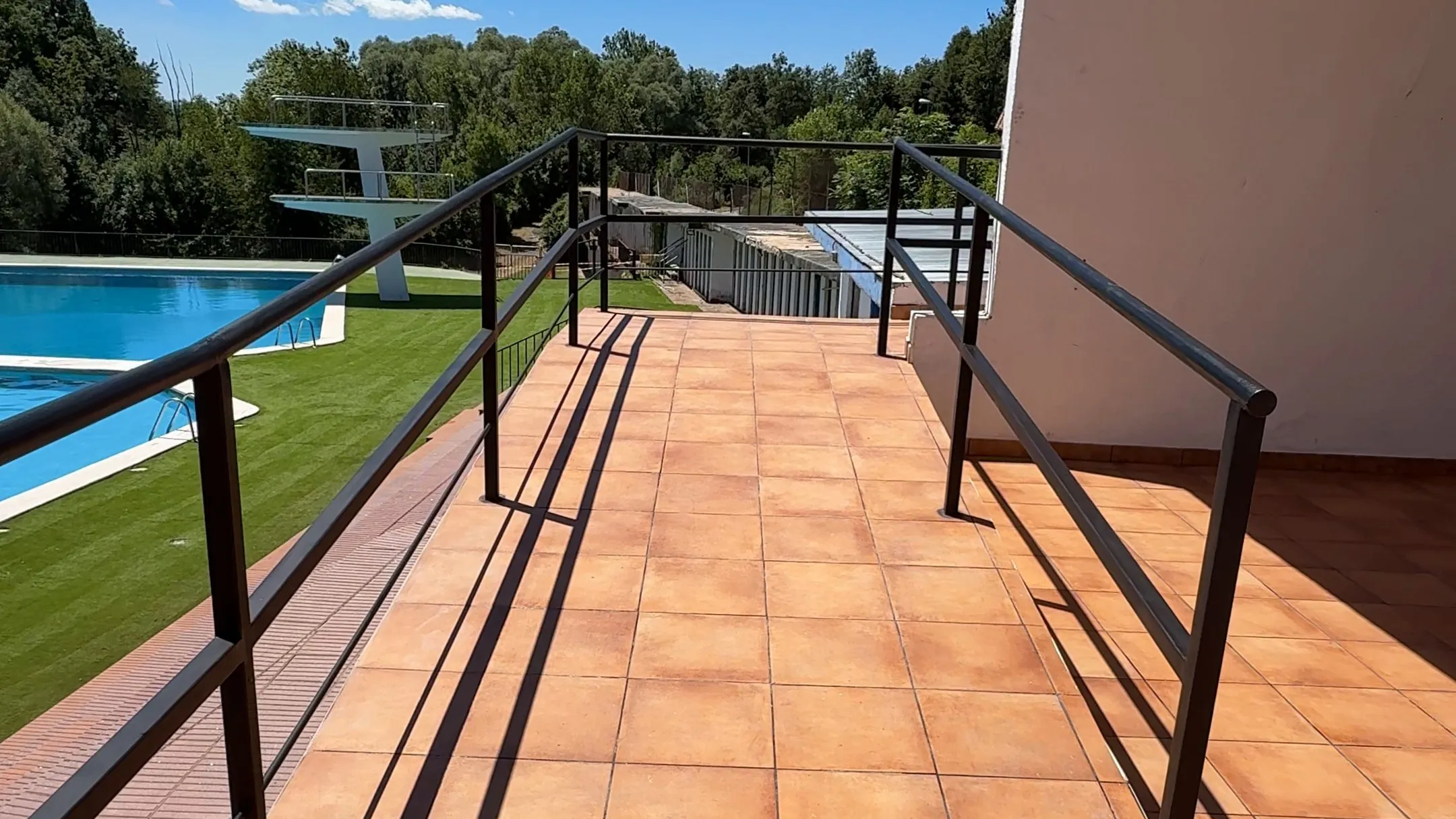 Rampa y terraza de baldosa cerámica de color terracota con barandillas metálicas negras a ambos lados, en la zona de la piscina de una comunidad de propietarios en Camprodon; al fondo, la piscina con trampolín, césped y árboles