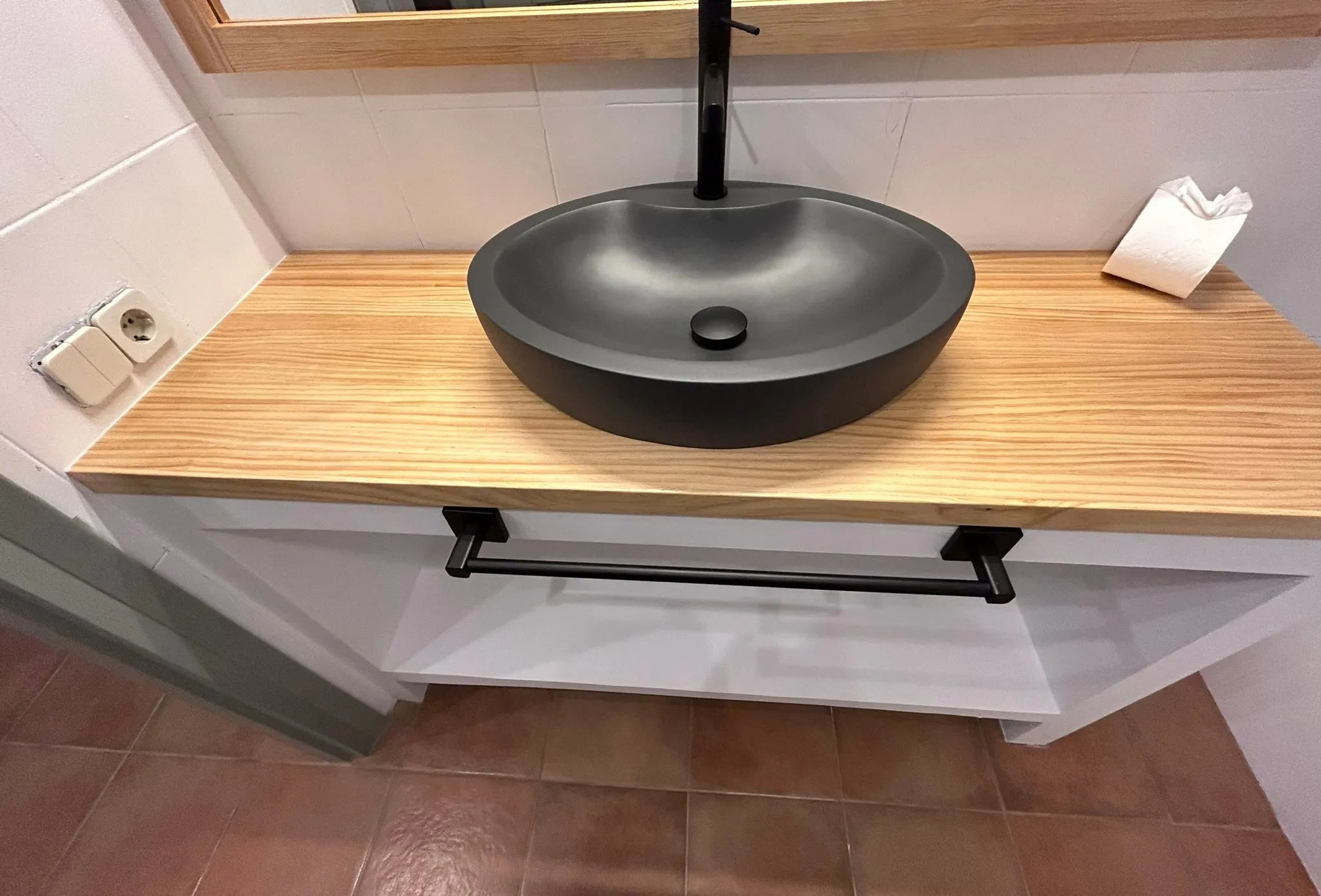Lavabo negro mate sobre una encimera de madera clara, con grifo negro alto; la base es blanca, con una balda abierta y un toallero negro, y el suelo es de baldosa de tono terracota