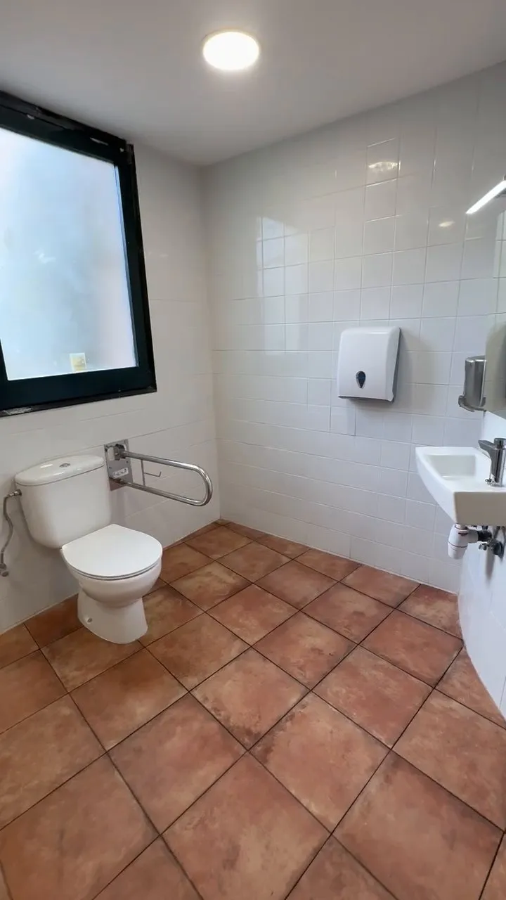 Un dels banys del Castell de Besalú reformat: parets enrajolades de blanc, terra de rajola terracota, finestra de marc negre, vàter blanc amb barra de suport abatible, lavabo petit de paret amb mirall i dispensador de tovalloles de paper