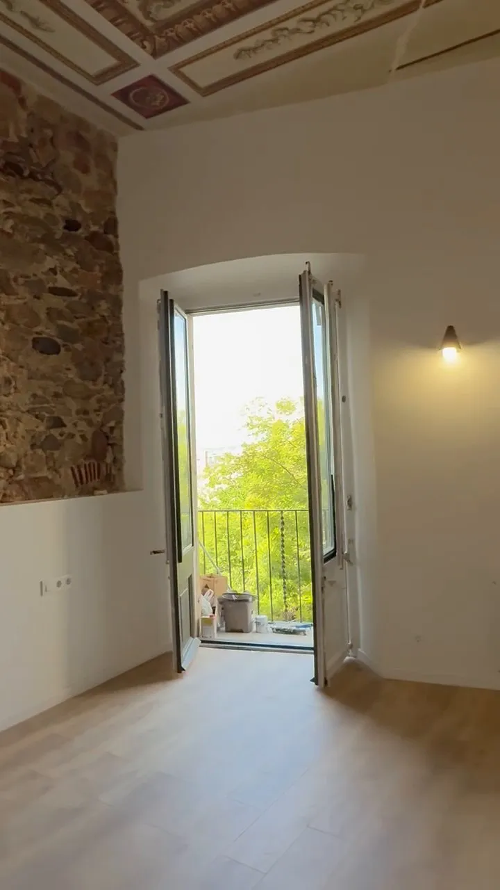 Estancia luminosa con balconera de dos hojas abierta a un balcón con barandilla metálica y cubos de obra, techo con molduras pintadas, muro de piedra a la izquierda y un aplique de pared