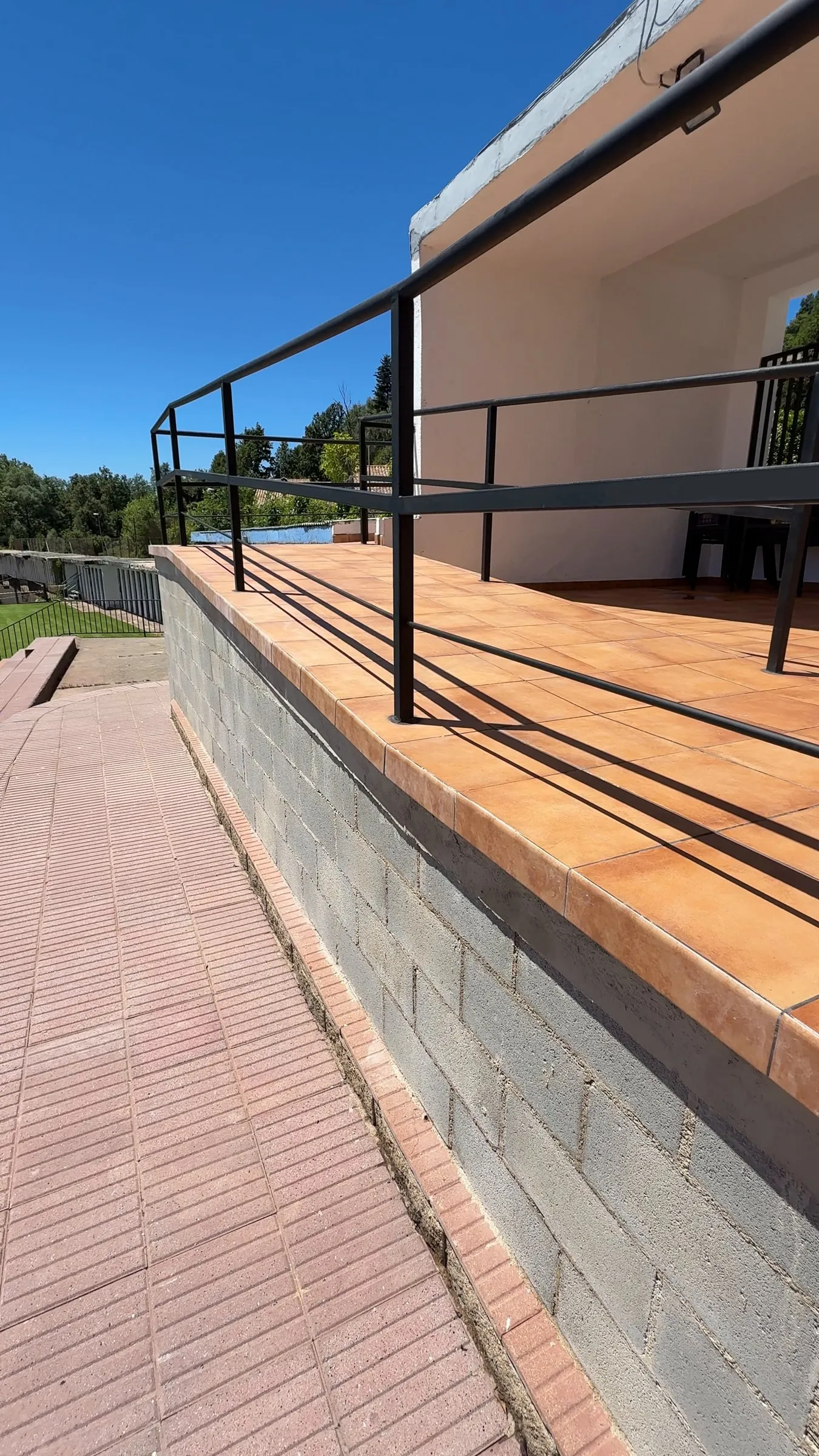 Terraza elevada con muro de bloque gris, borde rematado con baldosa cerámica y barandilla metálica negra, junto a un camino de pavimento rojizo; al fondo, el porche de un edificio blanco