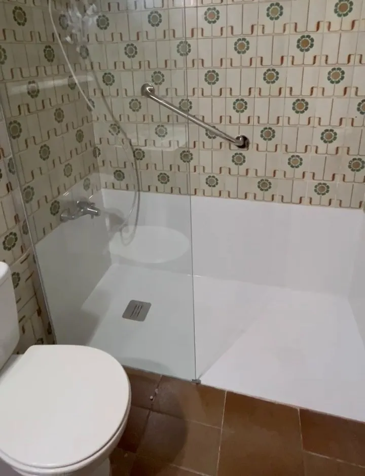 Plato de ducha blanco con desagüe de rejilla metálica, mampara fija de vidrio, grifo con flexo y barra de apoyo sobre el azulejo antiguo; abajo a la izquierda, el inodoro y el suelo marrón