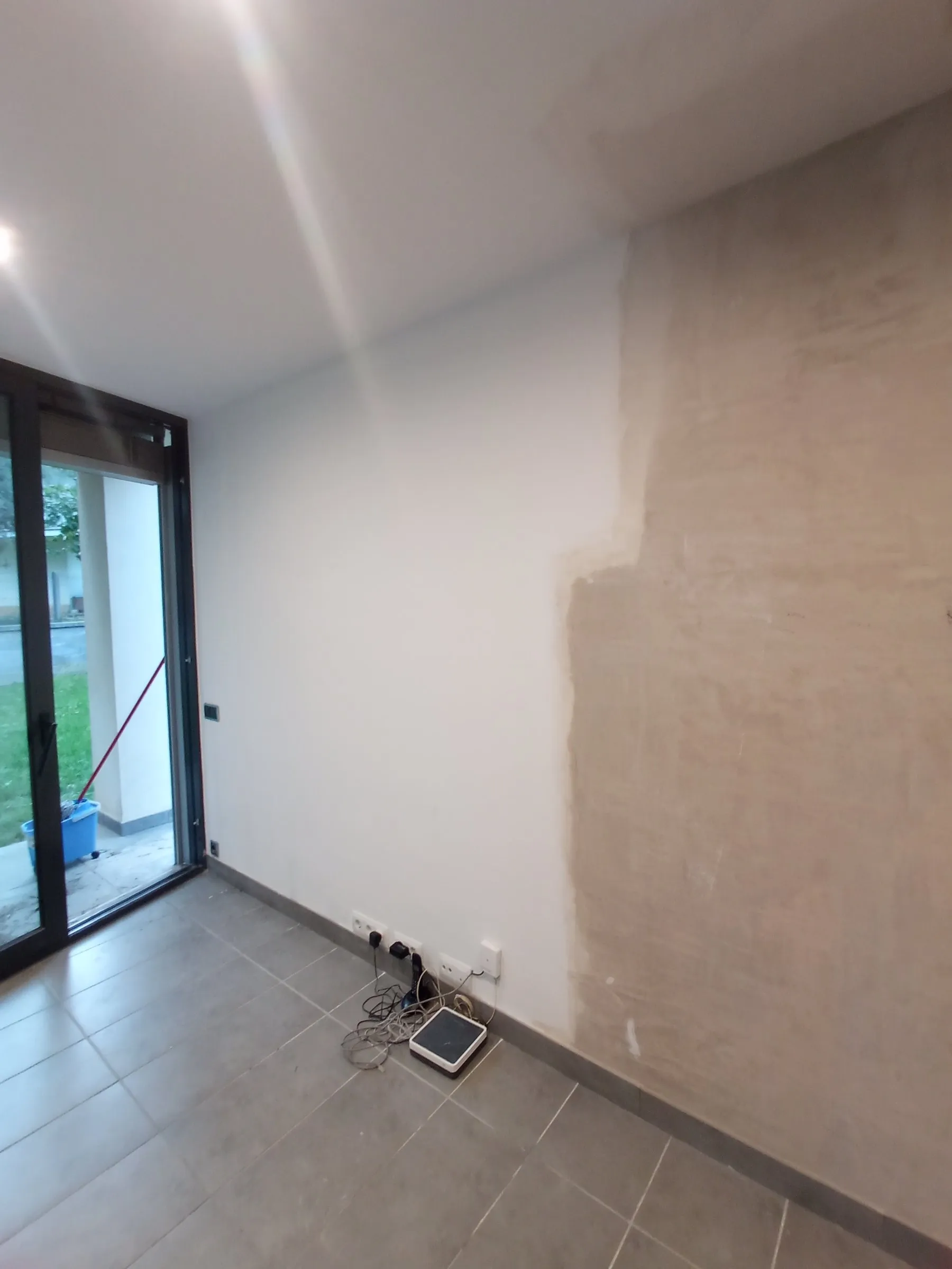 Salón de una vivienda en Cassà de la Selva con un tramo de pared enyesado y liso, listo para pintar, a la derecha, junto a una pared blanca; suelo de baldosa gris con rodapié gris y, a la izquierda, una puerta de cristal con marco negro que da a un jardín con césped