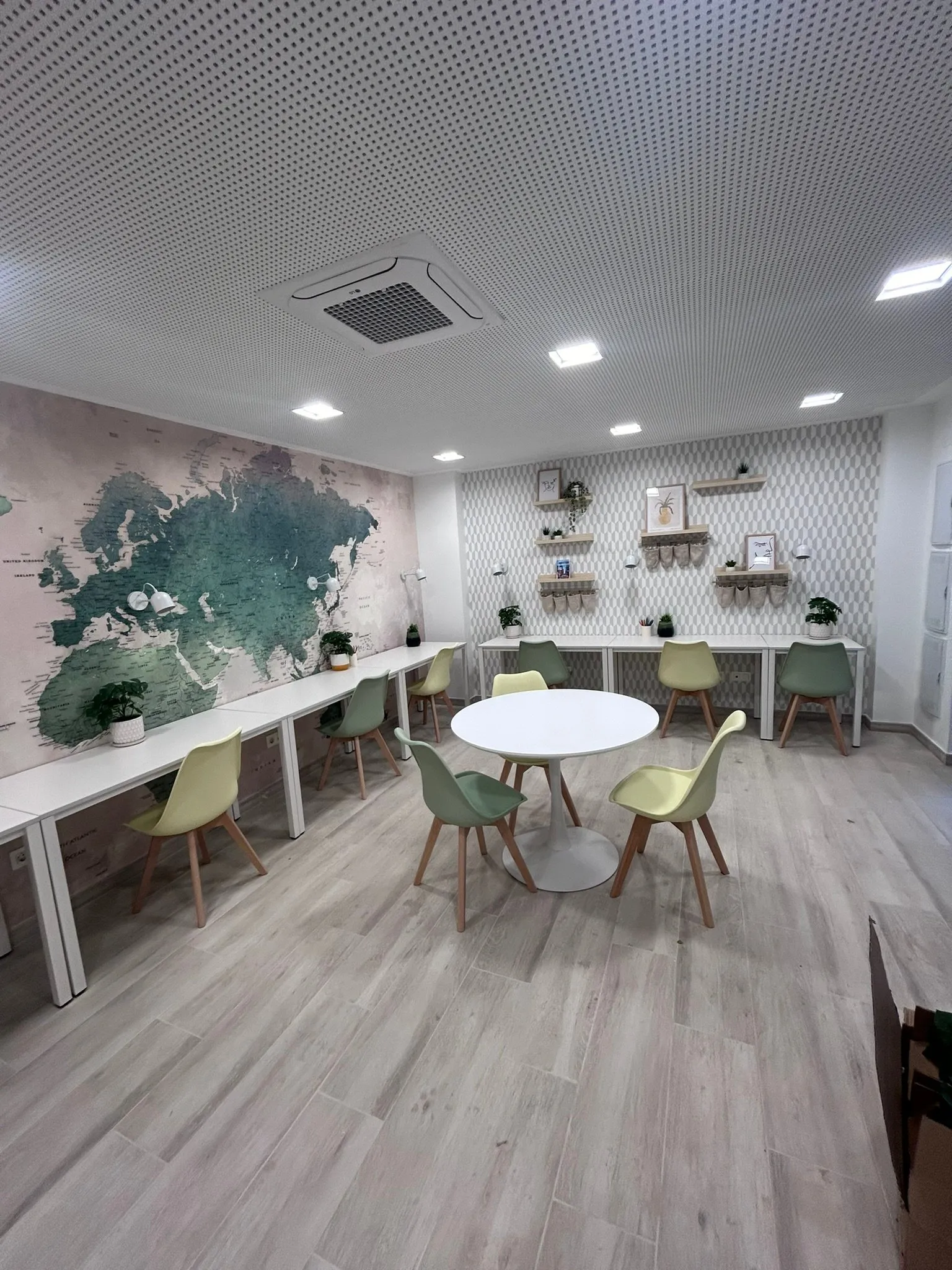 Sala amb taules llargues sota el mural de mapamundi, una taula rodona blanca amb cadires i prestatgeries amb quadres i plantes sobre paper pintat geomètric