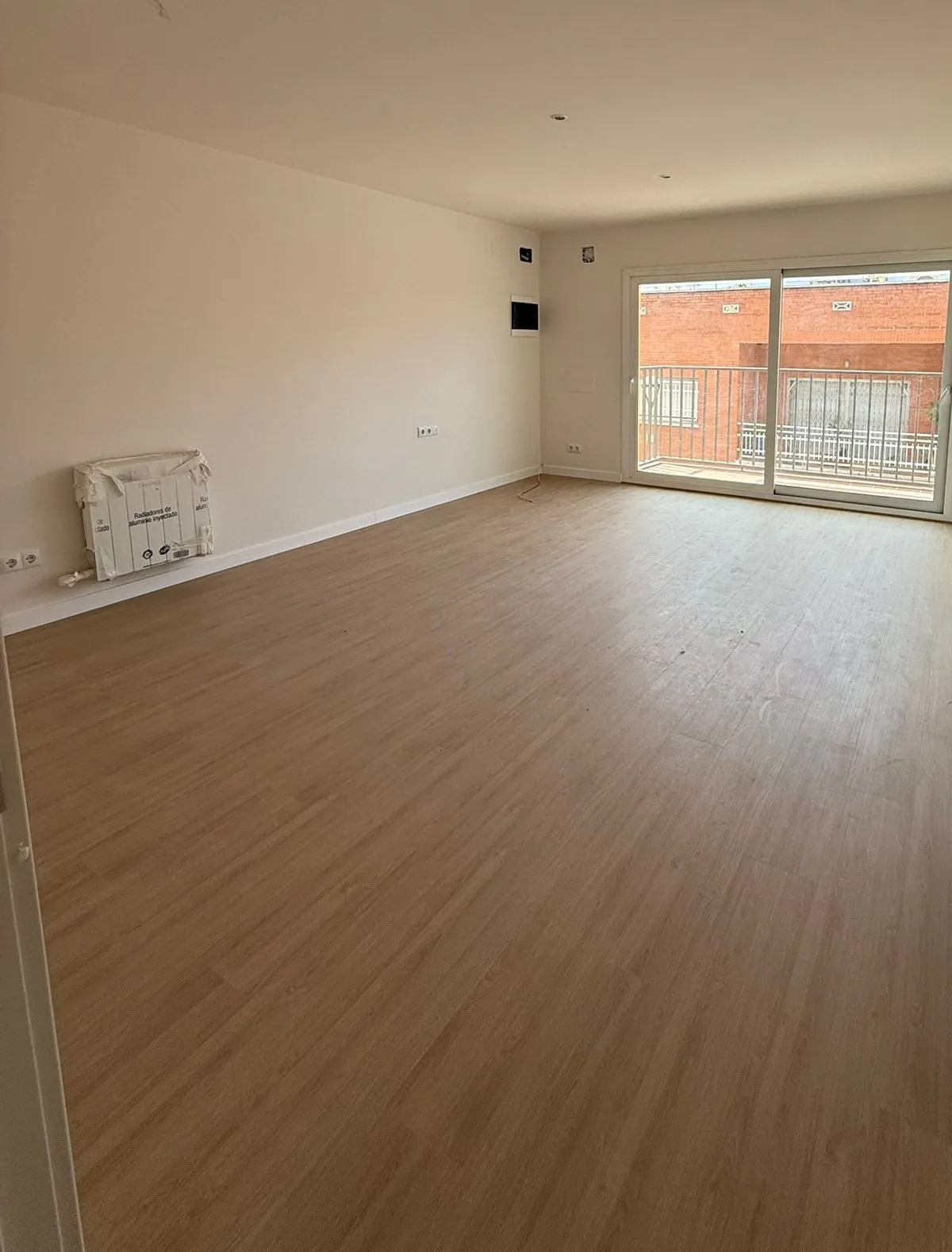 Salón con suelo de aspecto de madera clara, paredes blancas, un radiador aún embalado y una puerta corredera que da al balcón