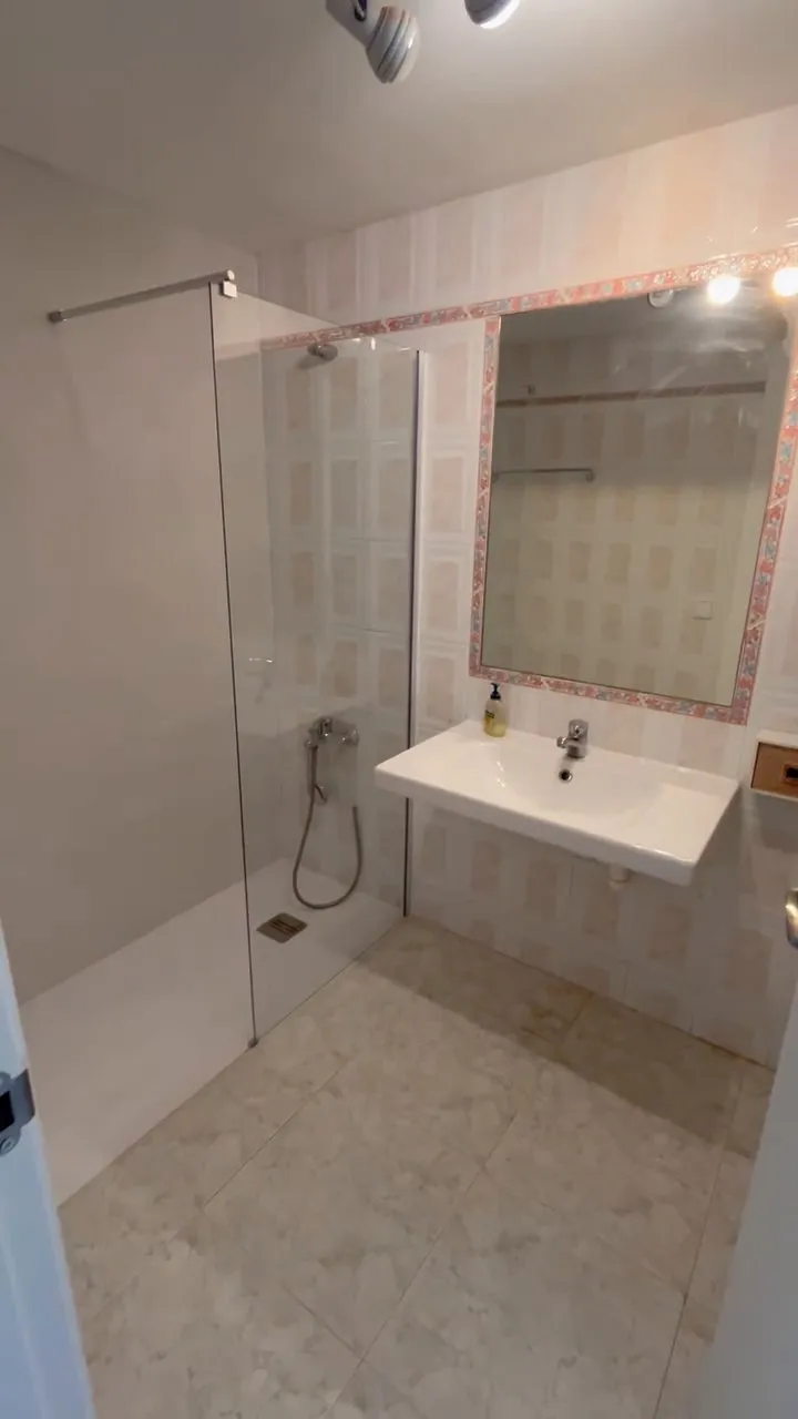 Vista general del bany amb la dutxa muntada: mampara fixa i plat blanc, i el lavabo sense moble sota el mirall amb sanefa de flors