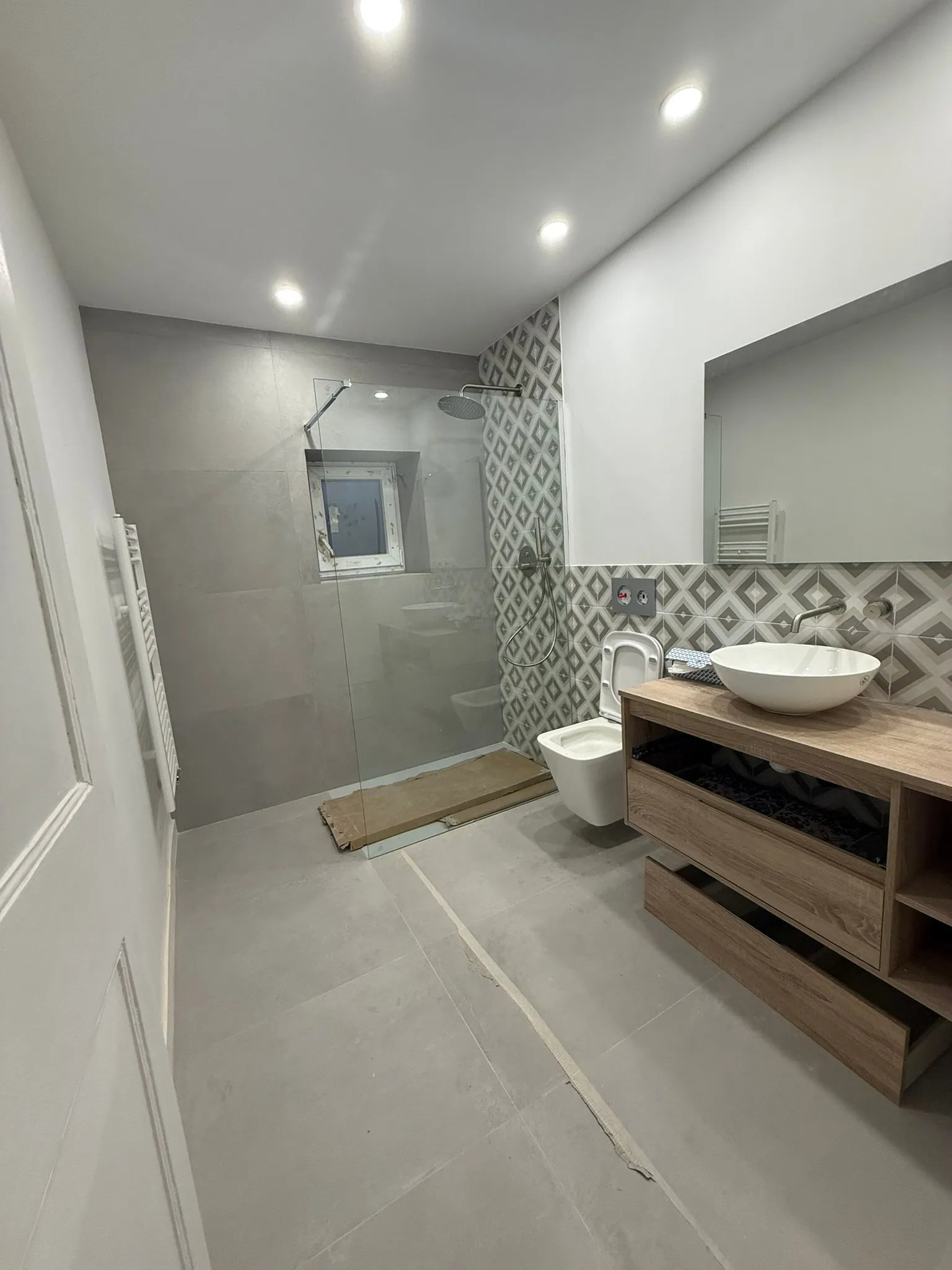 Baño con ducha de mampara de vidrio, baldosas grandes grises, revestimiento de rombos grises y blancos, lavabo tipo bol sobre encimera de madera con mueble suspendido con cajones, inodoro suspendido y espejo grande; quedan cartones de protección en la ducha