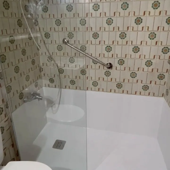 Detalle de la ducha: barra de apoyo metálica sobre el azulejo antiguo, grifo con flexo, plato blanco con desagüe de rejilla y mampara fija de vidrio transparente