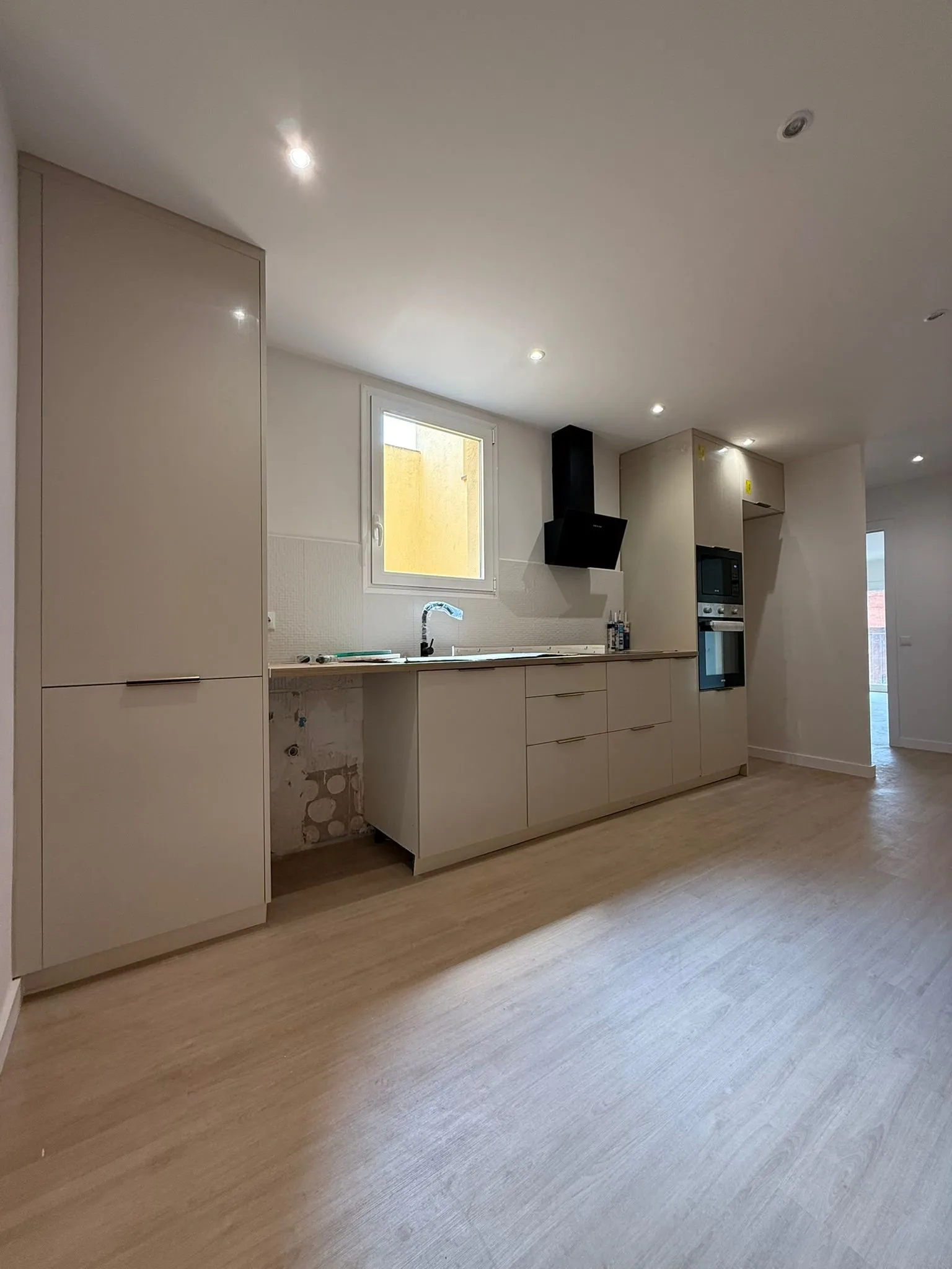 Cocina con muebles de frente beige, campana inclinada negra, horno y microondas en columna, encimera de acabado madera y suelo de aspecto de madera clara; bajo la encimera queda un hueco con la pared aún sin acabar