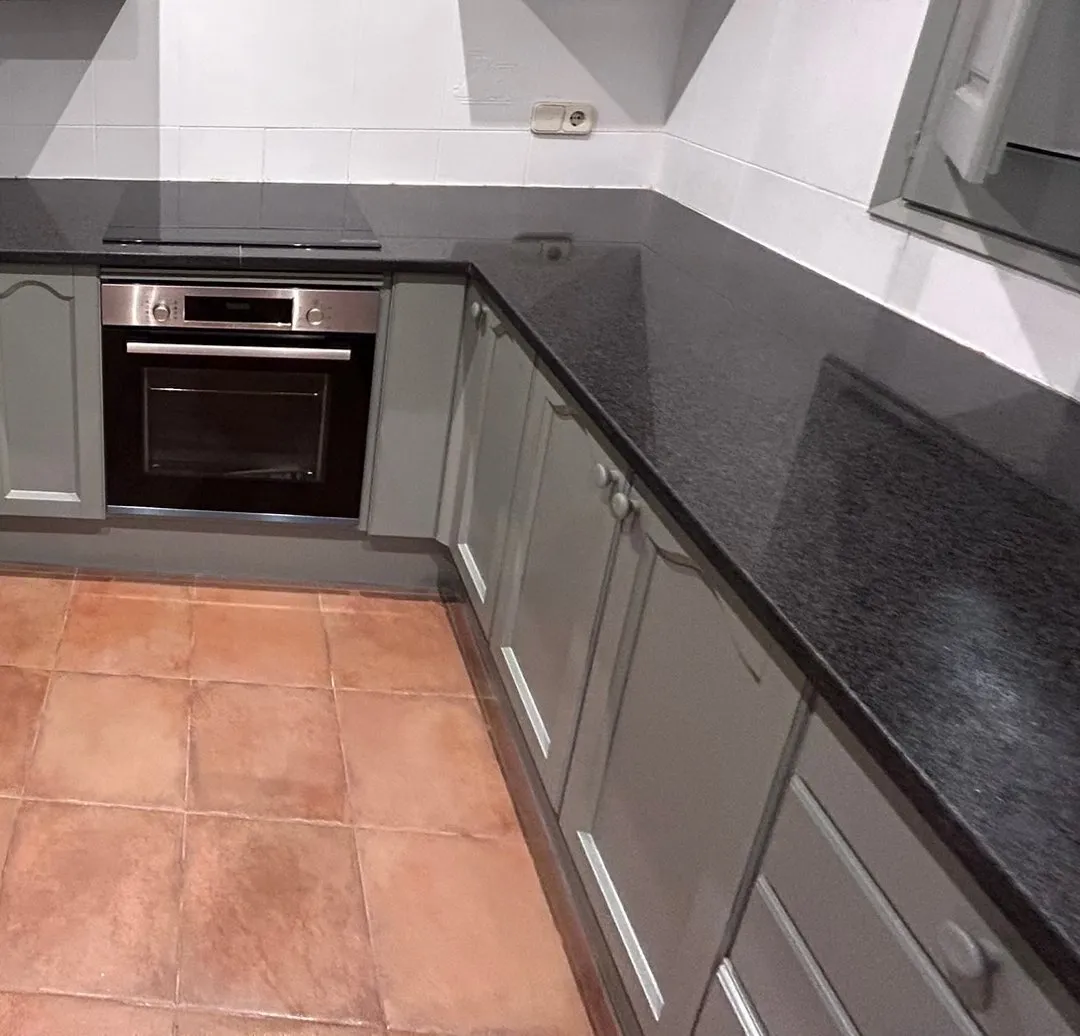 Cocina con muebles gris verdoso de puertas con moldura, encimera negra de aspecto de granito, horno empotrado de acero inoxidable con frontal negro, placa negra, pared de azulejo blanco y suelo de baldosa de tono terracota