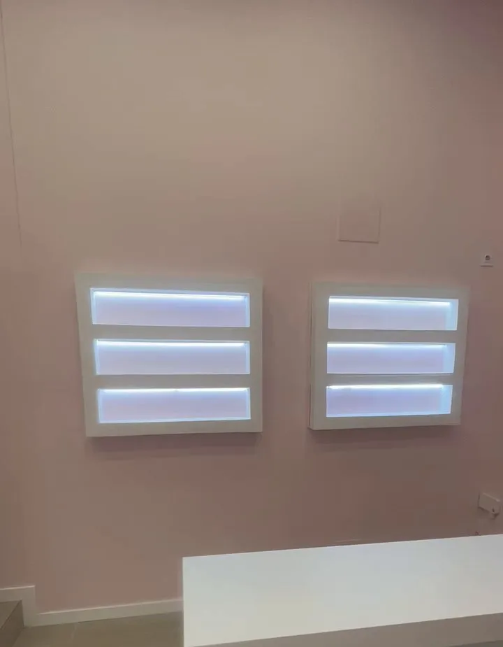 Paret rosa pàl·lid amb dos mòduls blancs de tres prestatges, il·luminats per dins amb tires LED de llum blanca; a baix es veu la vora d’una taula blanca