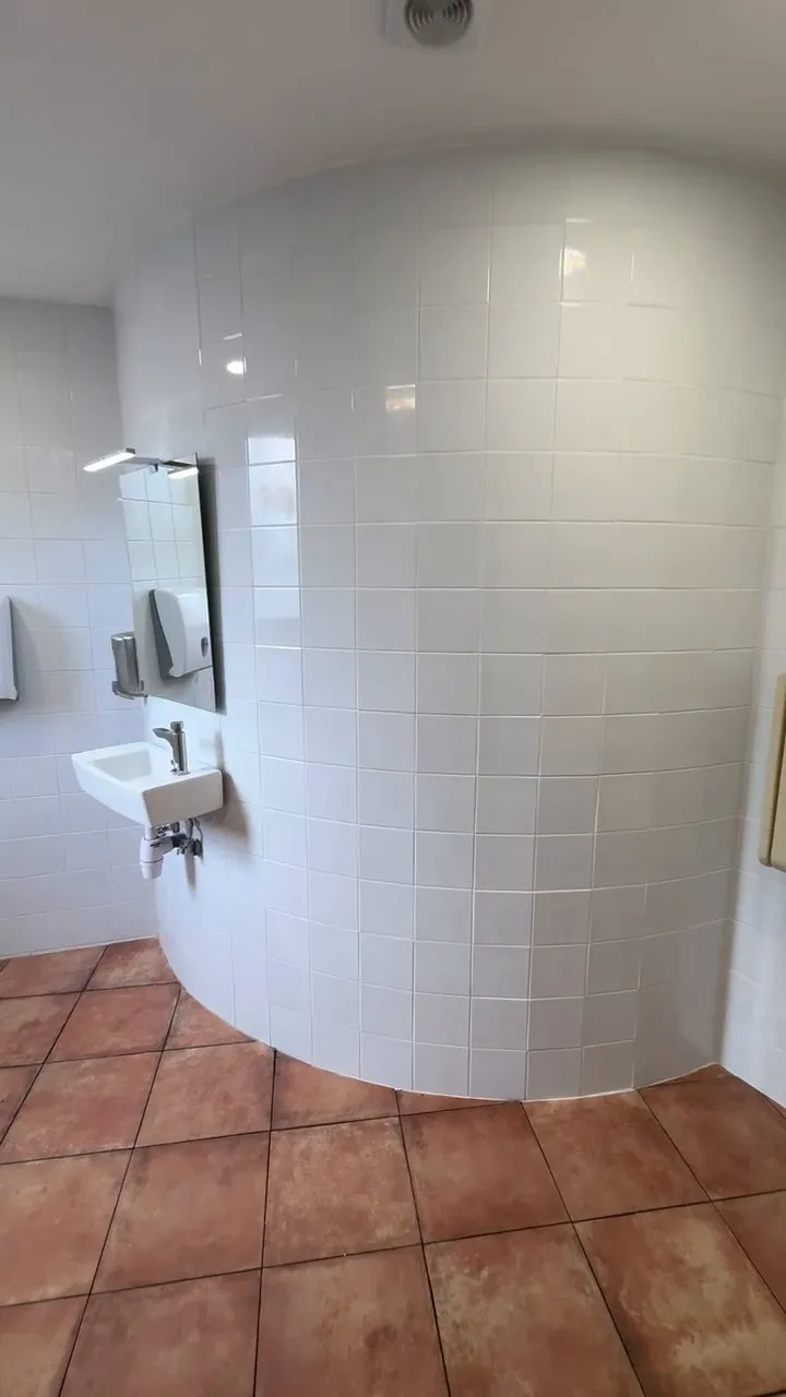 Paret corba enrajolada de blanc en un bany, amb un lavabo petit de paret i un mirall amb llum, sobre un terra de rajola terracota