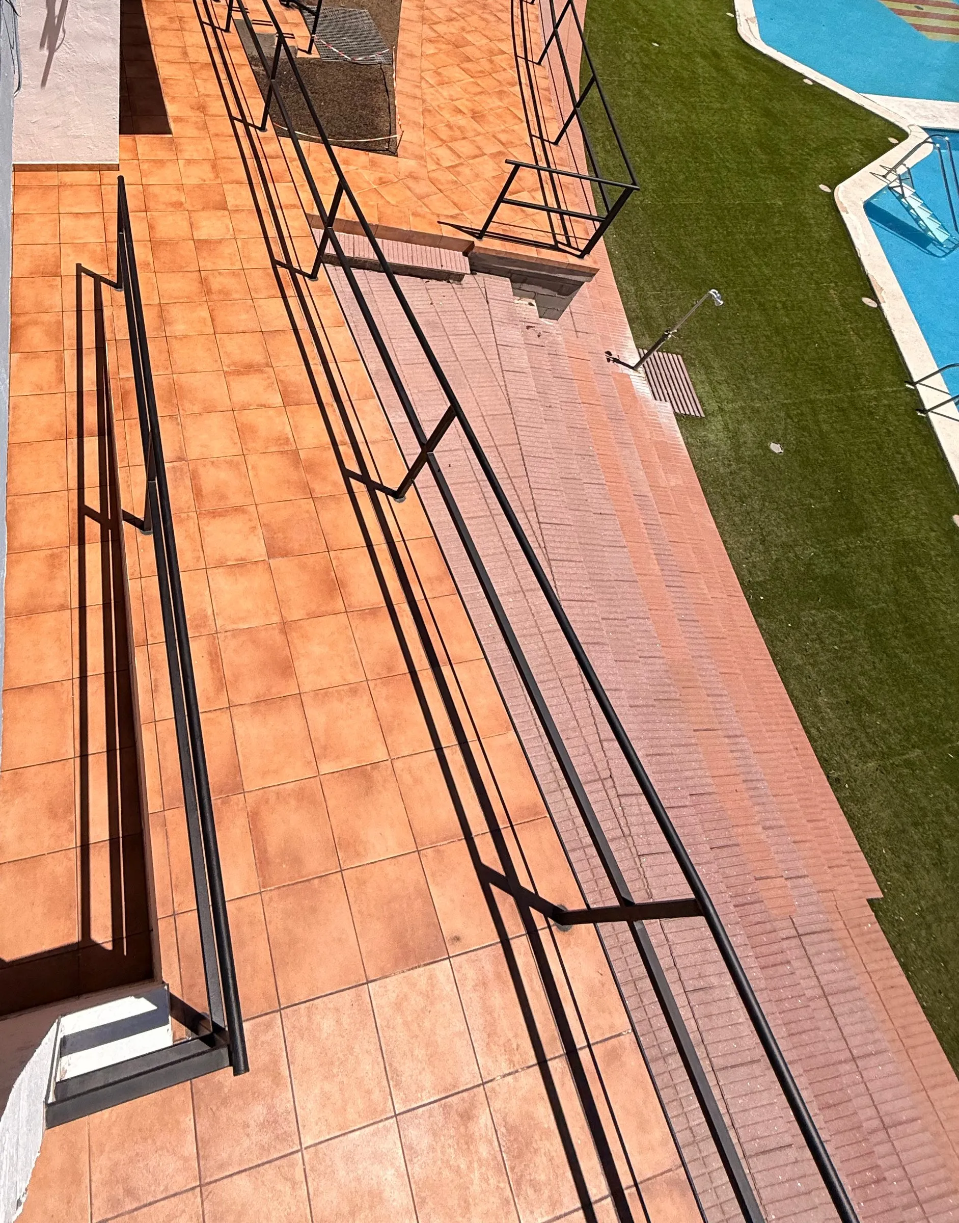 Vista desde arriba de una rampa de baldosa cerámica de color terracota con barandillas metálicas negras, junto a una grada de pavimento rojizo, con césped y una piscina a la derecha