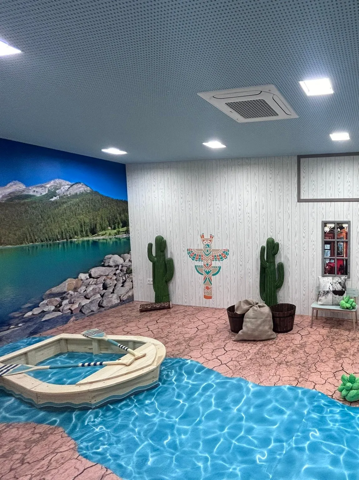 Sala de l’Oest, amb un mural d’un llac de muntanya, una barca de fusta amb rems sobre un terra decoratiu que imita un riu blau, cactus i un tòtem a la paret