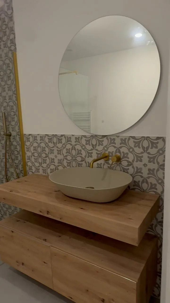 Lavabo ovalado color arena sobre encimera de madera con mueble suspendido, grifería dorada de pared, espejo redondo y baldosas con motivo floral gris; a la izquierda, la ducha con perfil dorado