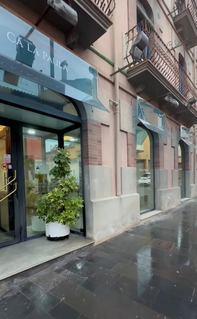 Façana de Ca la Paula a Castellfollit de la Roca: marquesina de vidre amb el rètol «Ca la Paula» sobre la porta vidriada, una planta en un test blanc al costat de l’entrada, balcons de ferro a la façana i la vorera mullada