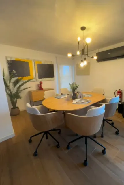 Sala de reuniones de la oficina de Platja d’Aro: mesa ovalada de madera clara con sillas giratorias tapizadas, lámpara de techo, dos cuadros abstractos y aire acondicionado en la pared