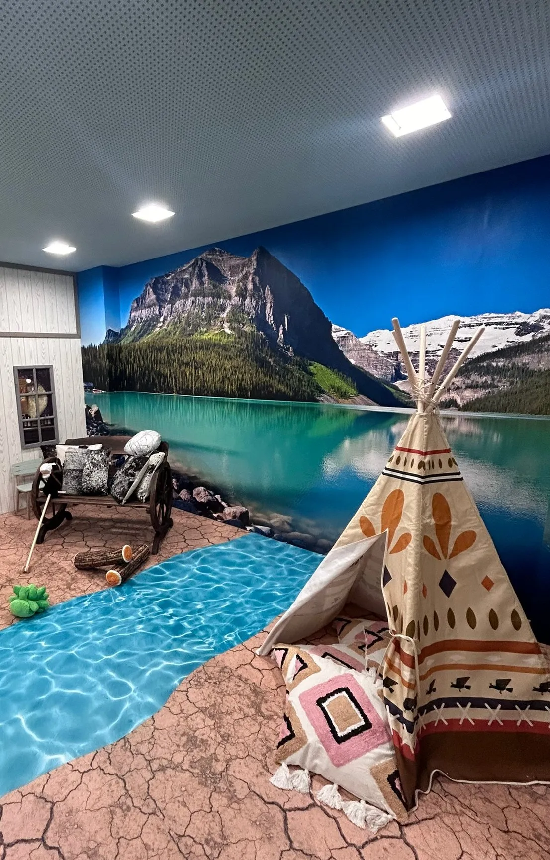 Racó de la sala de l’Oest amb un tipi al costat del mural d’un llac de muntanya, un banc rústic amb coixins i un riu blau sobre un terra decoratiu que imita terra esquerdada
