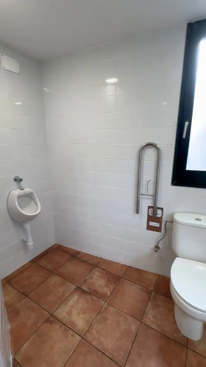 Racó d’un dels banys amb un urinari blanc de paret, una barra de suport en U d’acer, part del vàter i de la finestra de marc negre, parets enrajolades de blanc i terra de rajola terracota