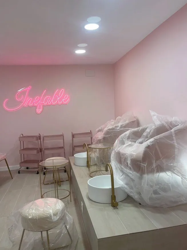 Sala del local d’Inefable a Figueres, amb parets rosa pàl·lid i el neó «Inefable» encès a la paret del fons; al davant, una plataforma d’acabat clar amb dos lavabos rodons blancs i aixeteria daurada, butaques cobertes amb plàstic, carretons roses, una tauleta auxiliar daurada i un puf, sota focus encastats al sostre