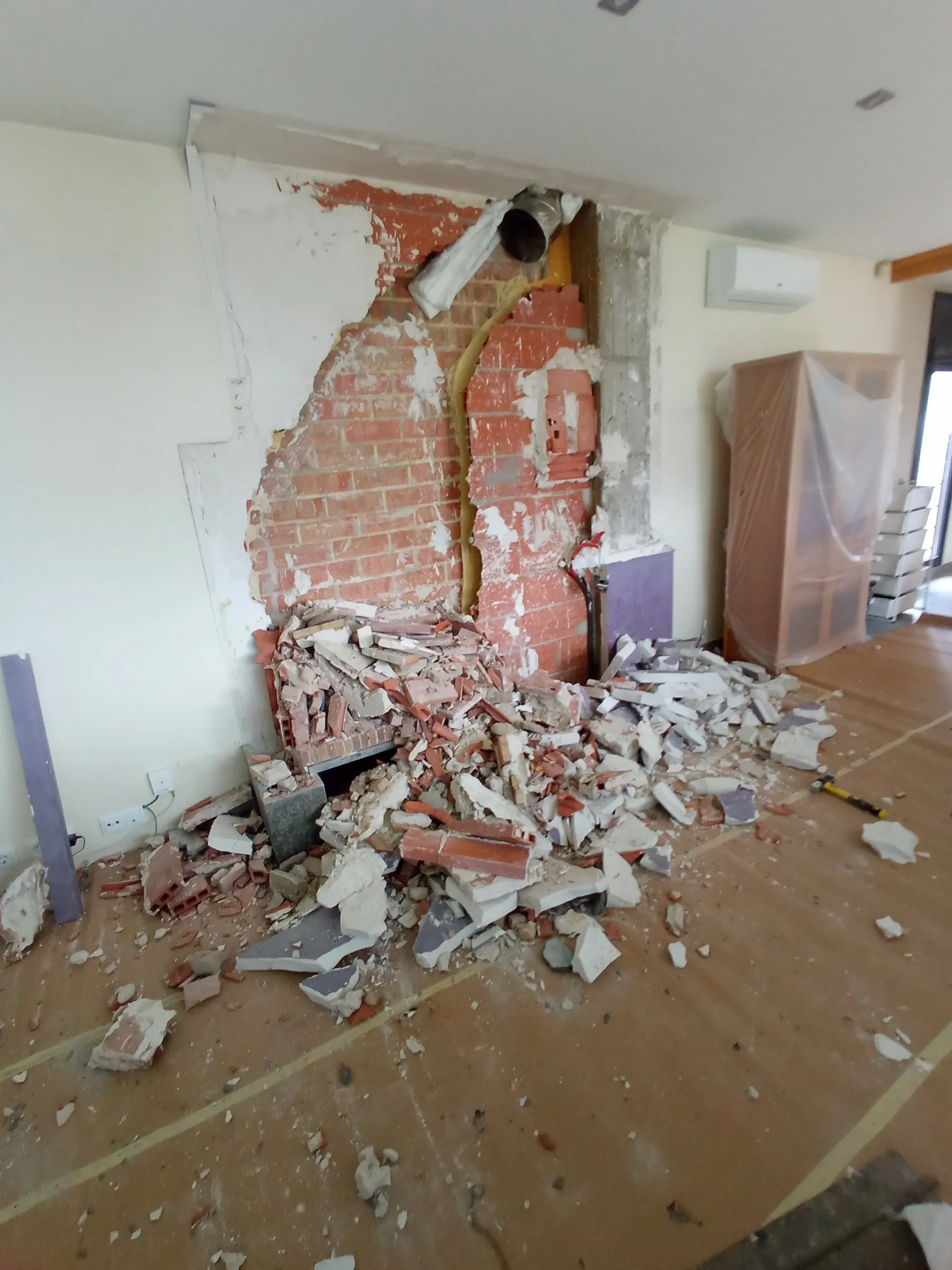 Pared de ladrillo a la vista tras derribar la chimenea, con un montón de escombro de ladrillo, yeso y trozos de revestimiento lila sobre el suelo protegido con cartón; arriba asoma un tubo metálico