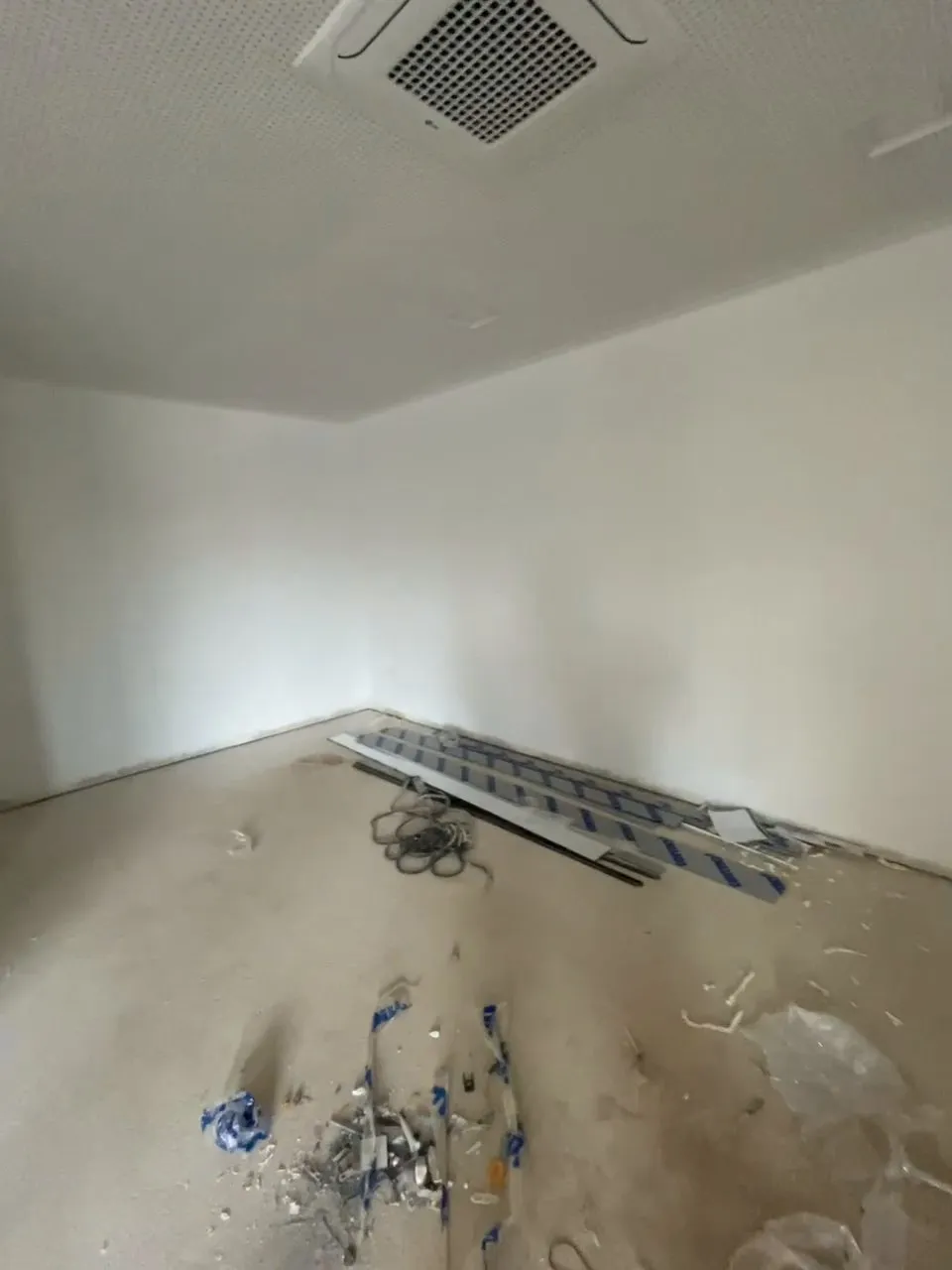 La sala de jocs buida, en obres: parets blanques, fals sostre perforat amb climatitzador de cassette, terra sense acabar amb restes d’obra i material recolzat a la paret