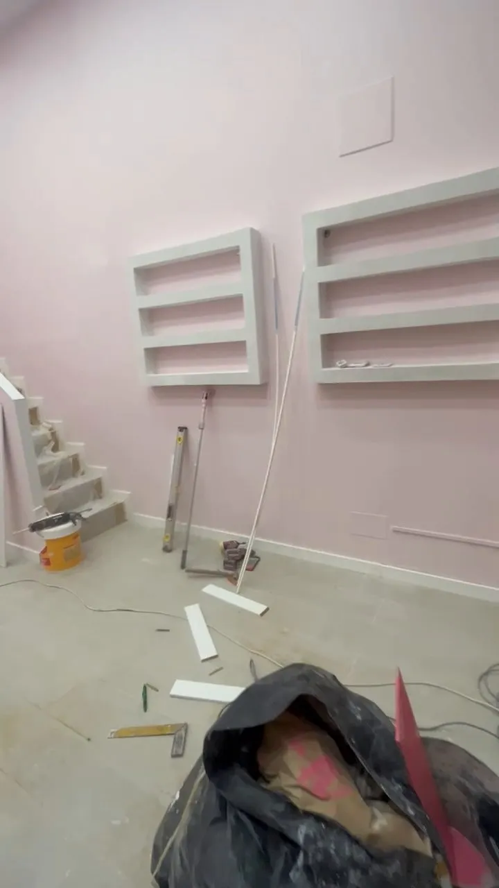Sala en obres amb la paret pintada de rosa pàl·lid i dos mòduls de prestatges blancs de tres lleixes; a terra hi ha eines, cables, tires blanques soltes i un sac negre amb cartró, i a l’esquerra es veu una escala amb els graons coberts